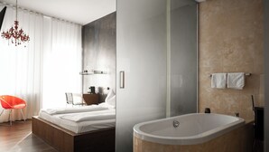A modern bedroom with a bed, bedside table, and a red chair. It also features a large bathtub and a shower area. - ImperialArt (Merano)