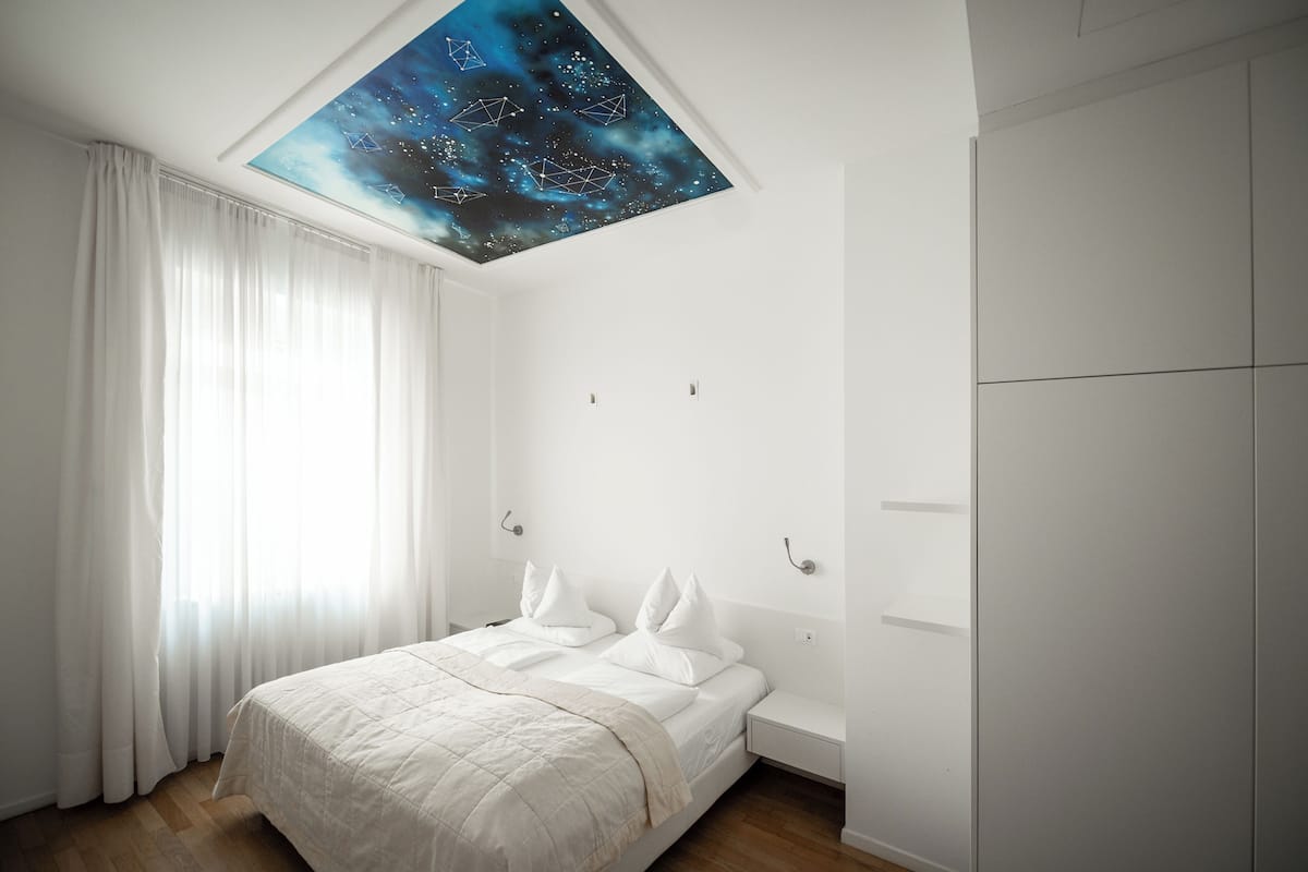 Un dormitorio con cama, cortinas blancas, una mesita de noche, un armario y un tragaluz en el techo con diseño de noche estrellada.