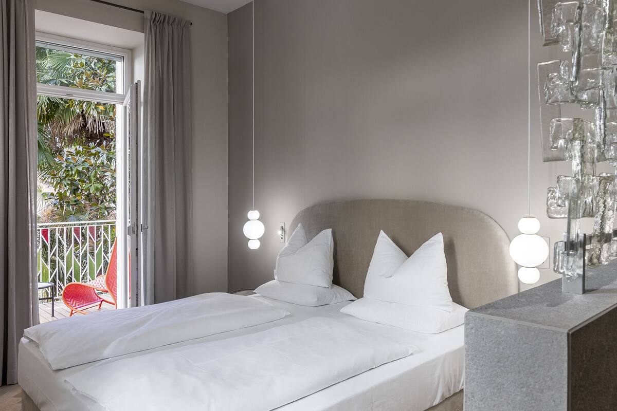 Un dormitorio con cama, ropa de cama blanca, cabecera gris y un balcón con barandilla y sillas.