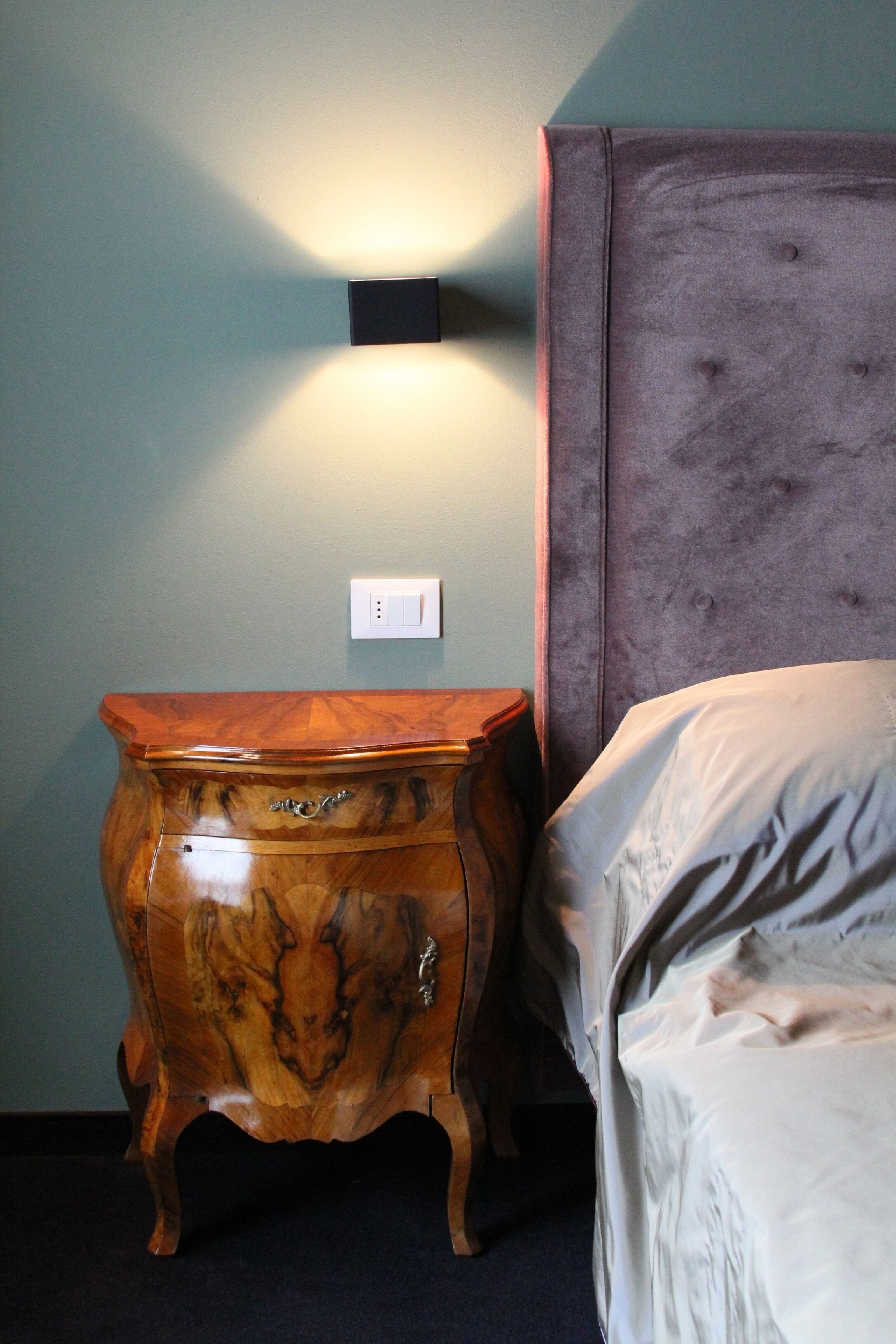 Un dormitorio con mesita de noche de madera, cama con cabecero e iluminación fijada a la pared.