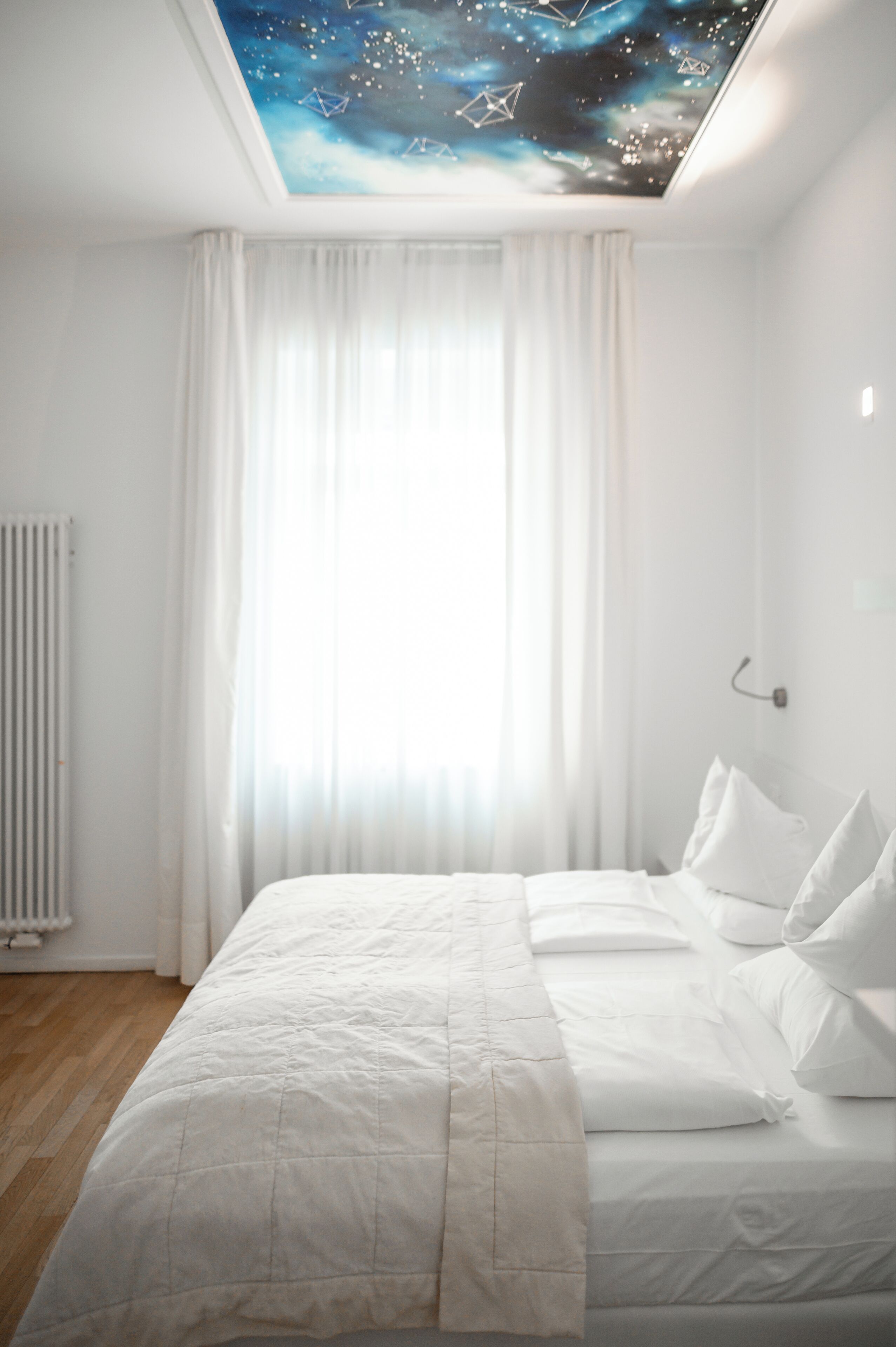 A bedroom with a bed, white curtains, a radiator, and a starry ceiling design.