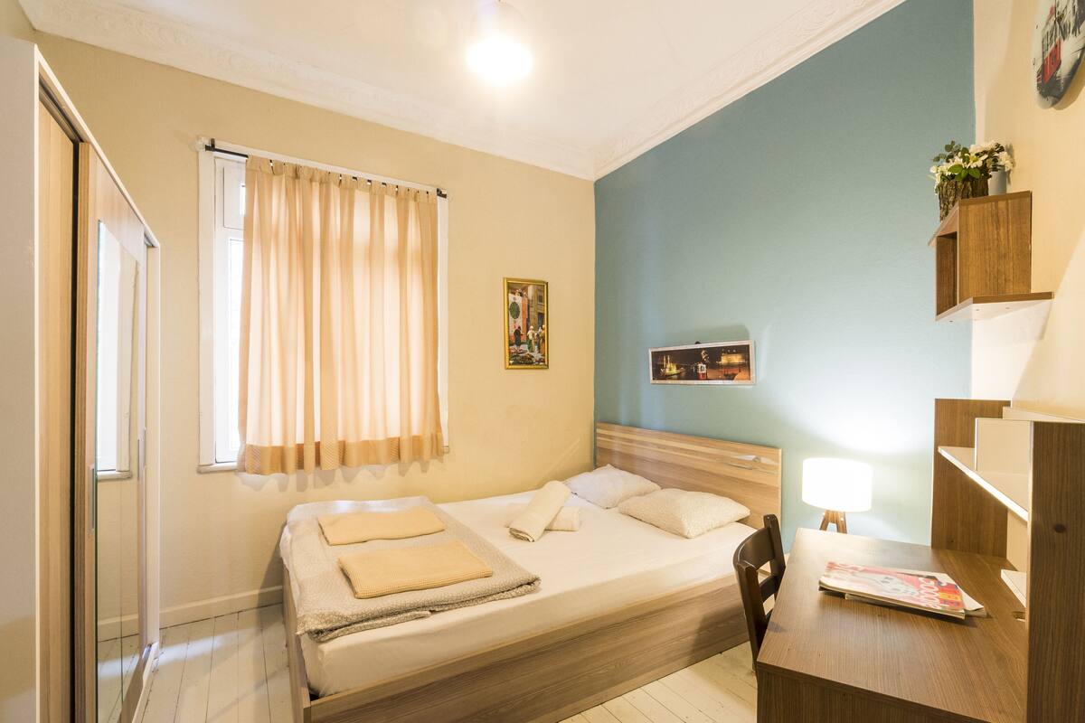 Une chambre à coucher comprenant un lit, un bureau, une chaise, une fenêtre avec des rideaux, une étagère fixée au mur et un tableau.