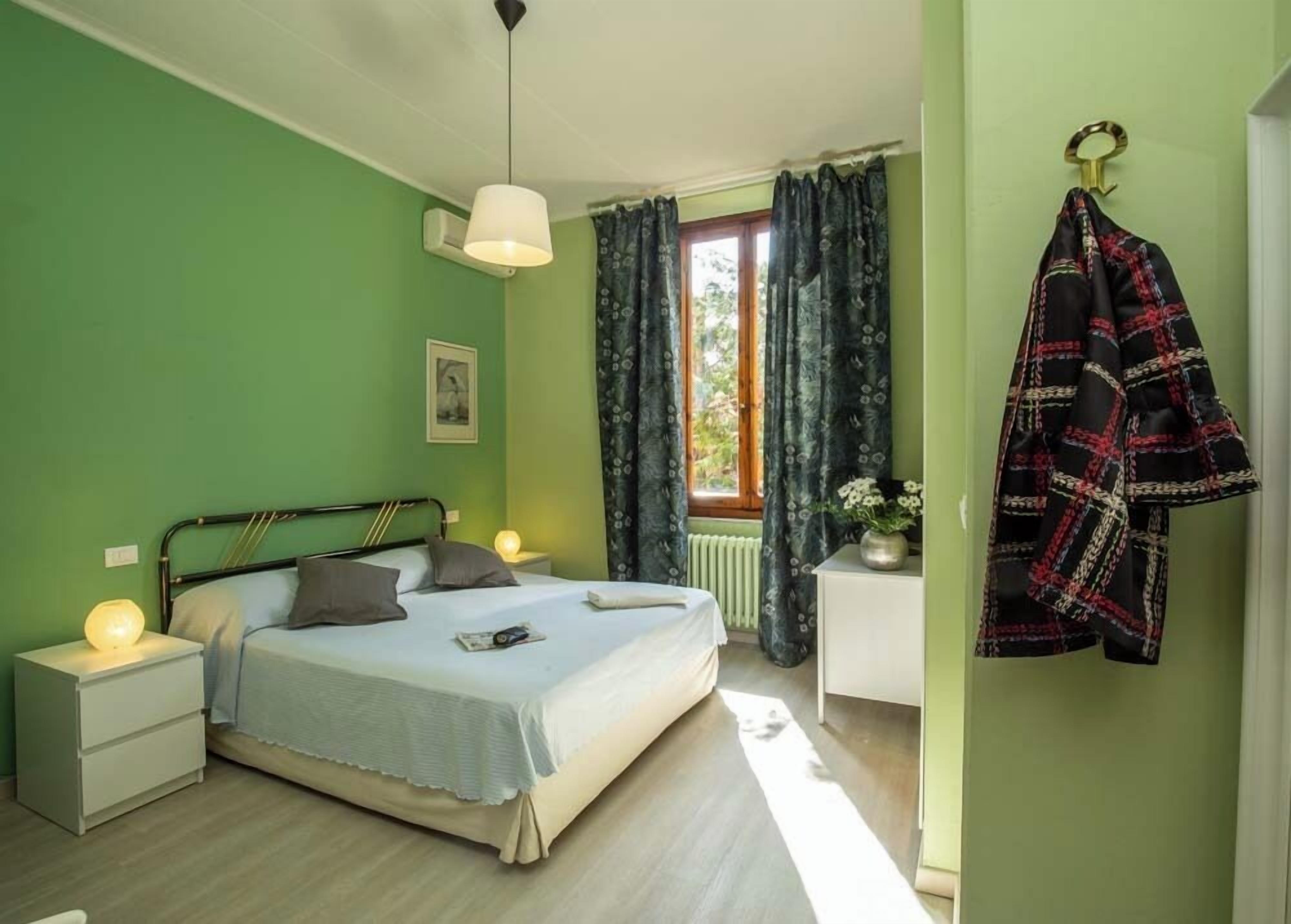 Un dormitorio con cama, lámparas de noche, ventana con cortinas, radiador y abrigo colgado en la pared.