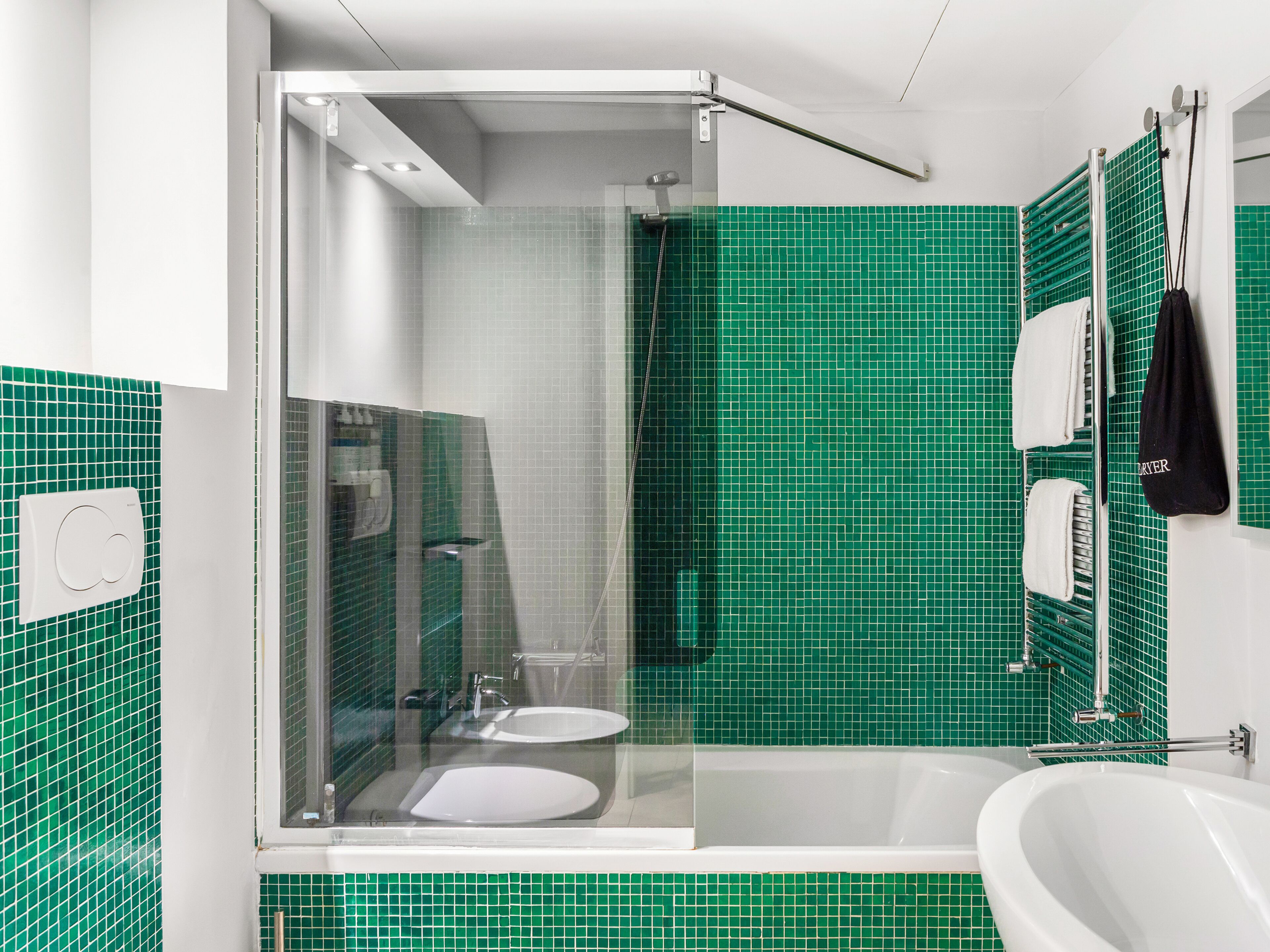 A modern bathroom with green mosaic tiles, a glass-enclosed shower, a white sink, and a bathtub.