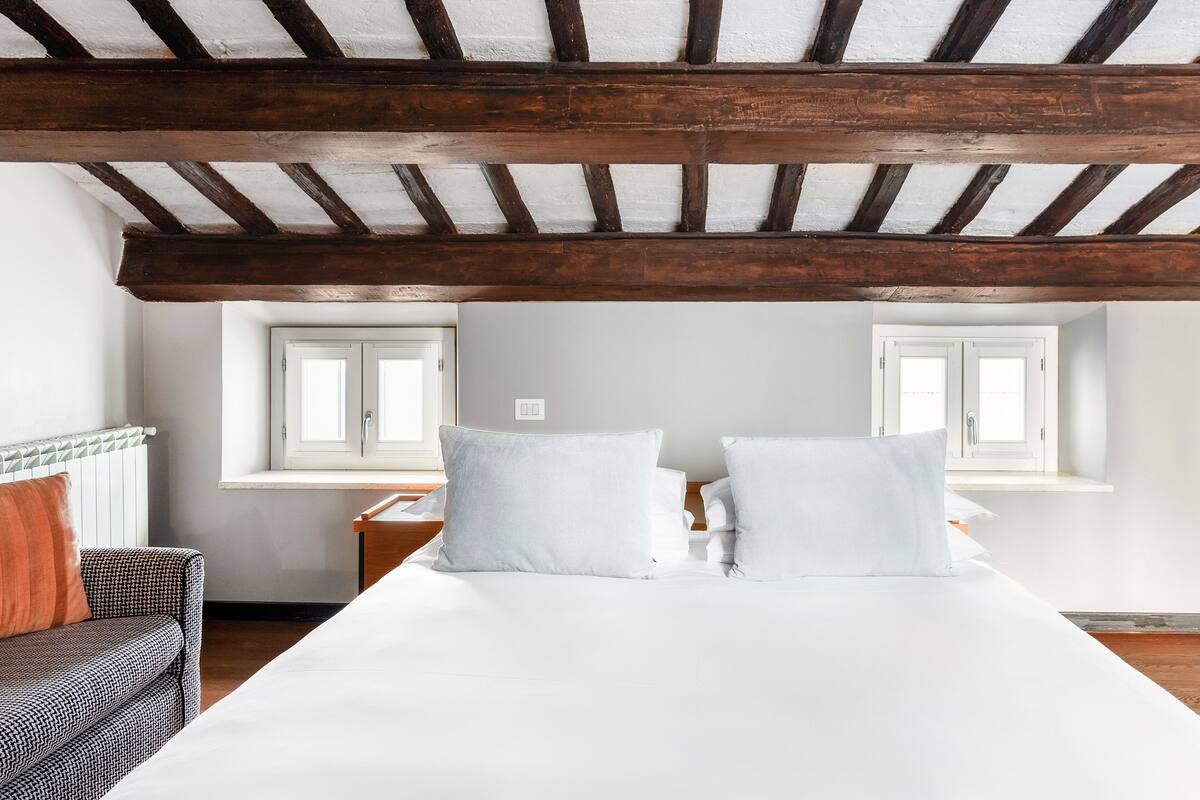 Une chambre avec un grand lit, des poutres en bois au plafond, deux fenêtres, une table de chevet et une chaise.