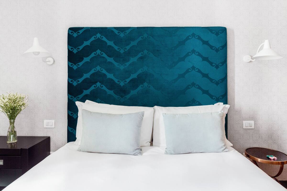 Un lit bien fait, avec une tête de lit à motifs turquoise, deux oreillers et un vase de fleurs posé sur une table de chevet.