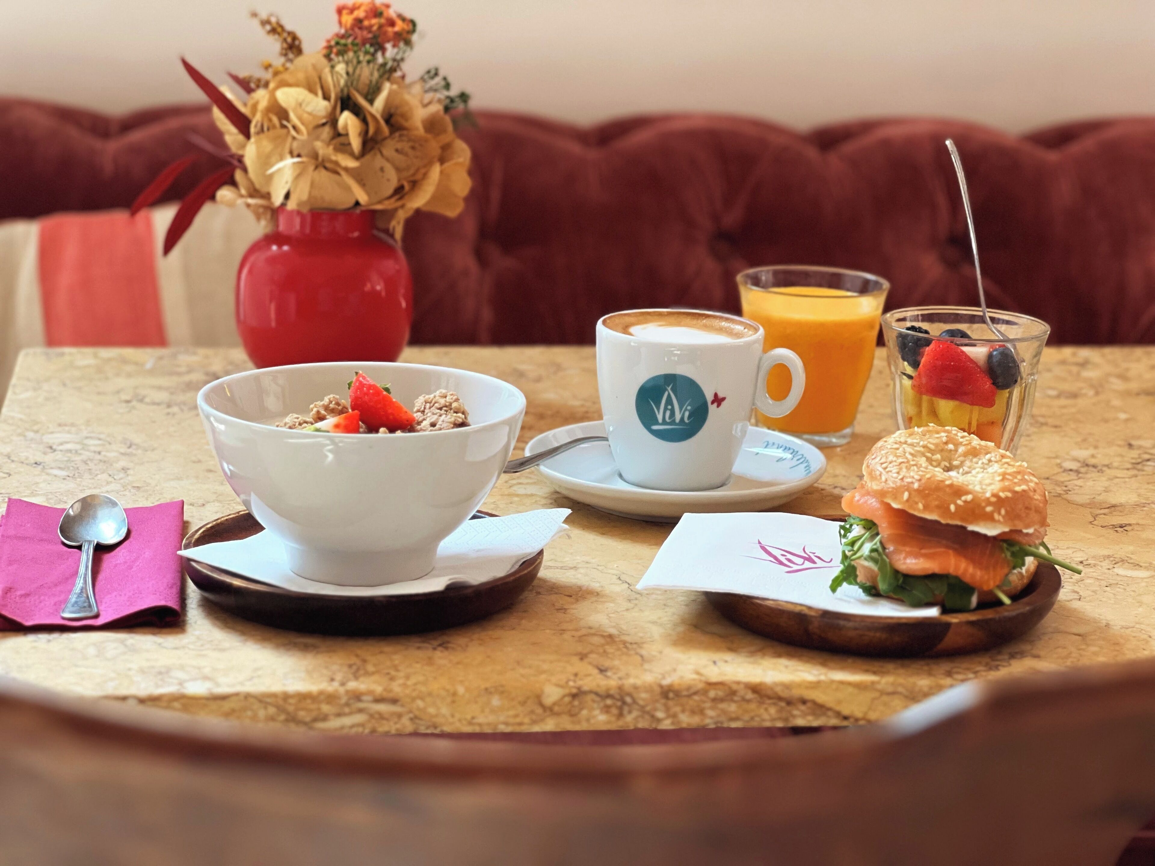 A breakfast setting with a bowl of cereal, a cup of coffee, a glass of orange juice, a sandwich, and a vase of flowers on a table.