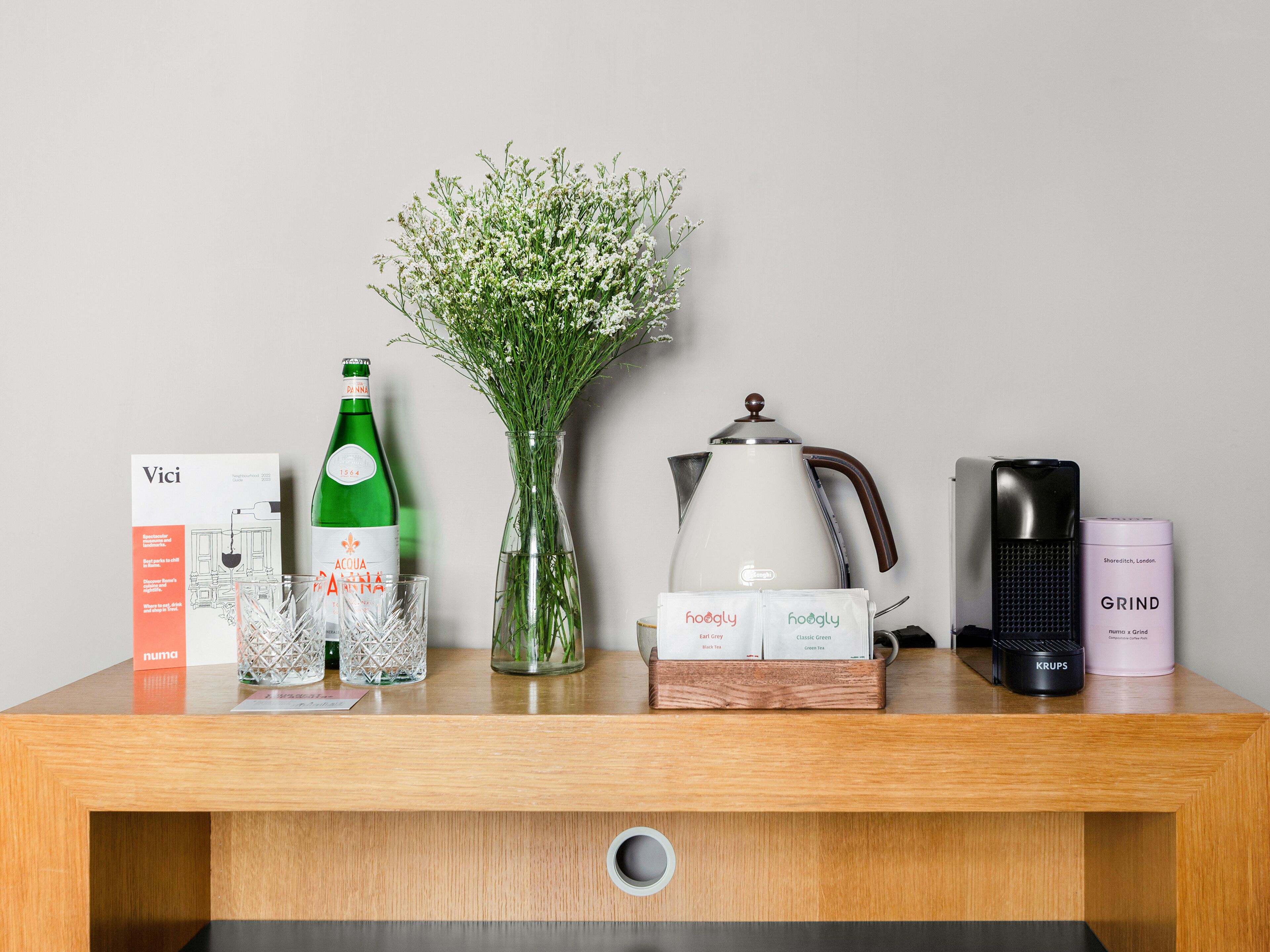 A wooden table with a bottle of water, a vase of flowers, a coffee maker, and a box of tea.