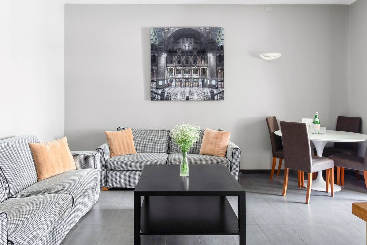 Un salon moderne avec une table basse noire, un canapé à carreaux et un coin repas comprenant une table blanche et des chaises marron.