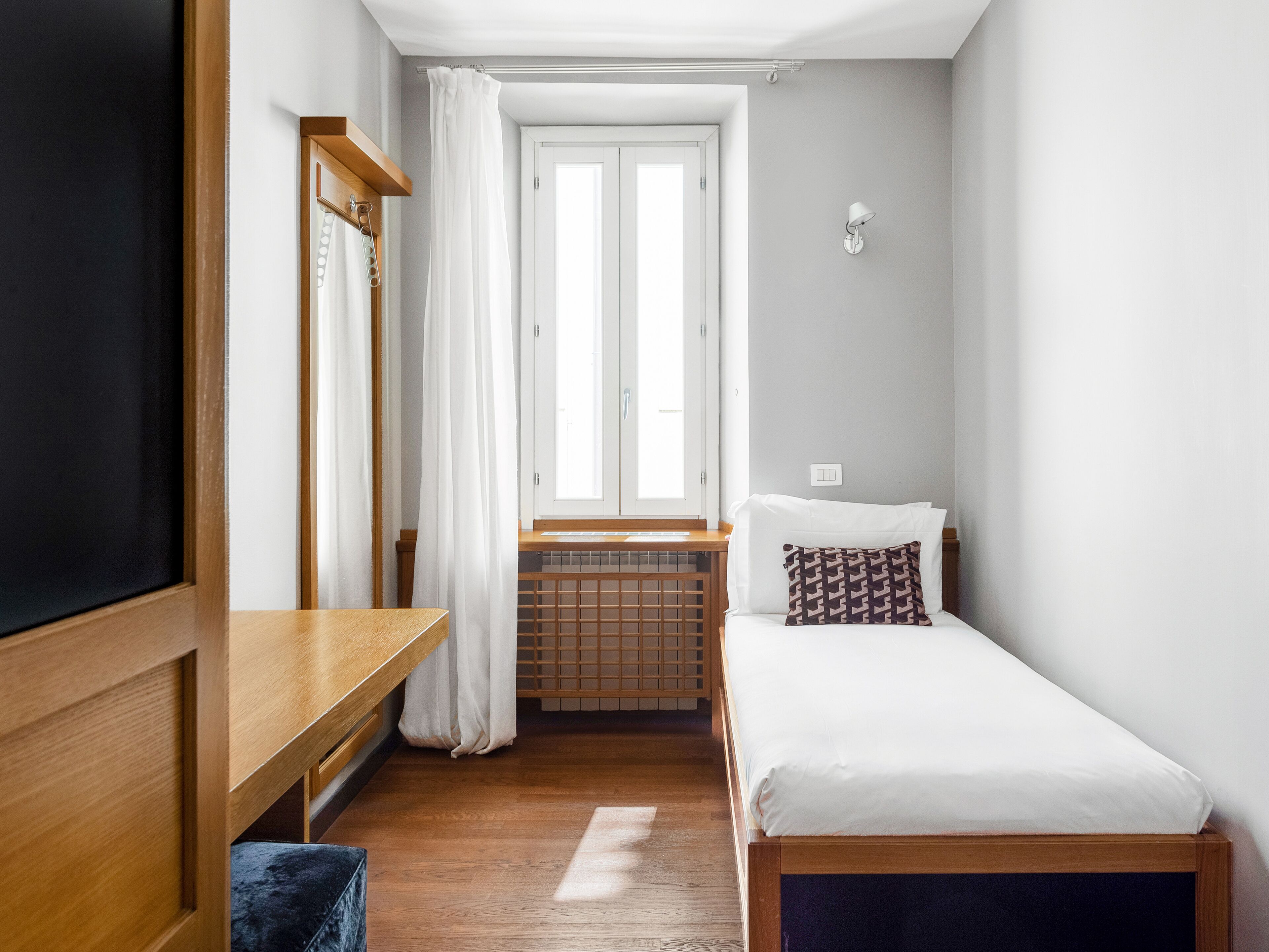 A small, well-lit bedroom with a single bed, a wooden desk, and a radiator.