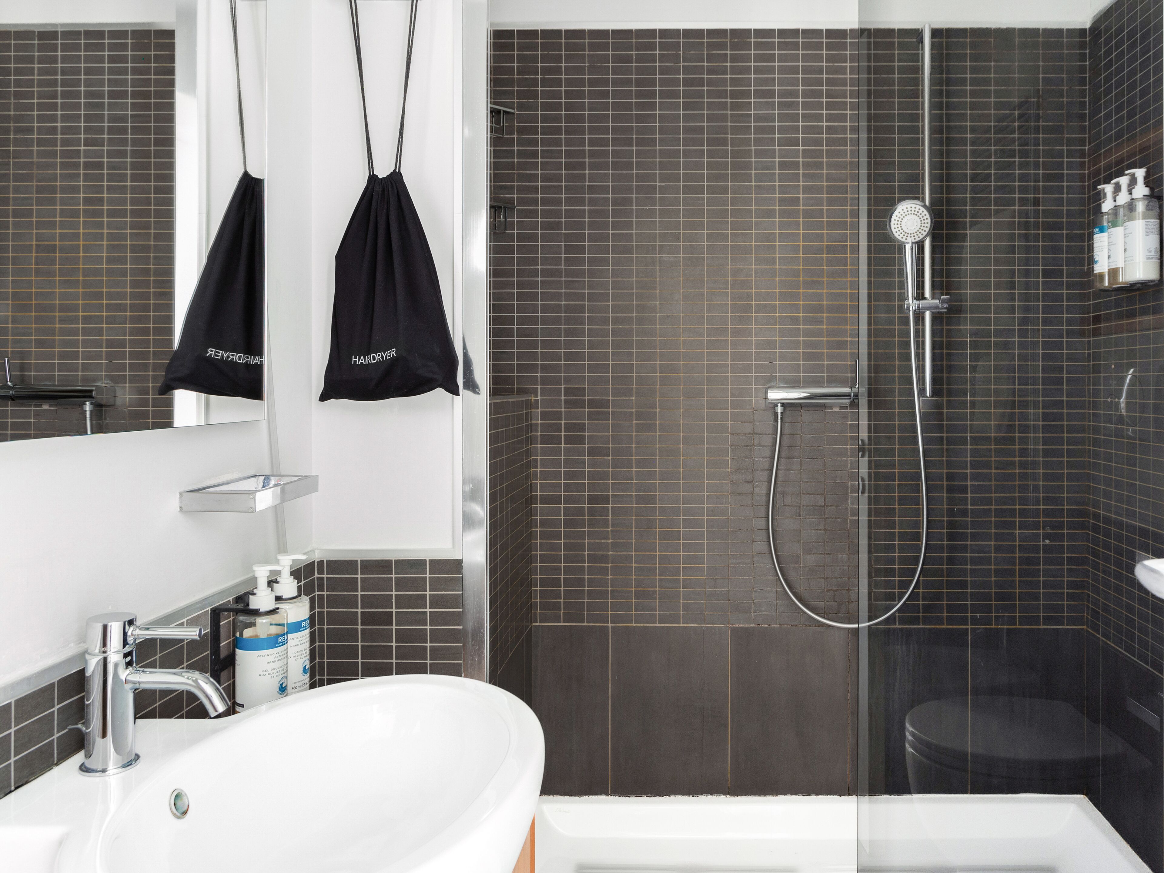 A modern bathroom with a white sink, a glass shower enclosure, and dark tiled walls.