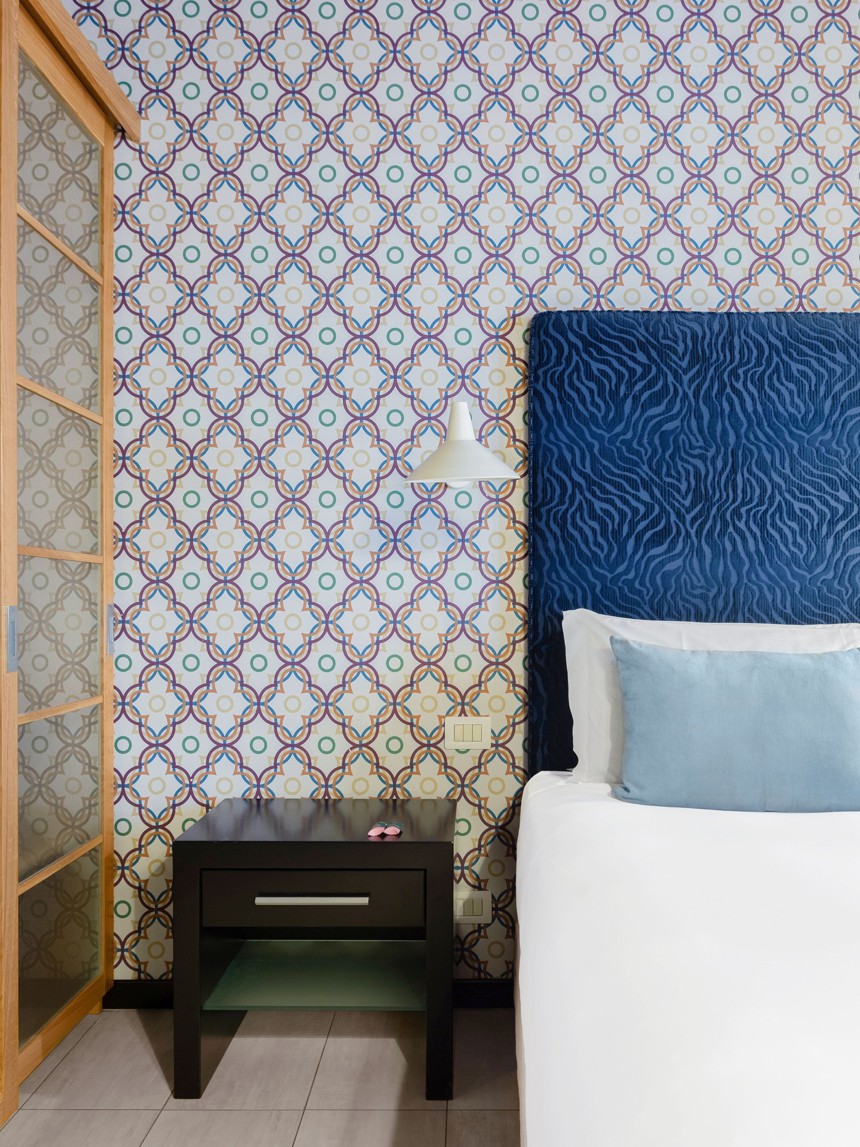 A bedroom with a patterned wallpaper, a blue headboard, a bedside table with a lamp, and a bed with blue pillows.