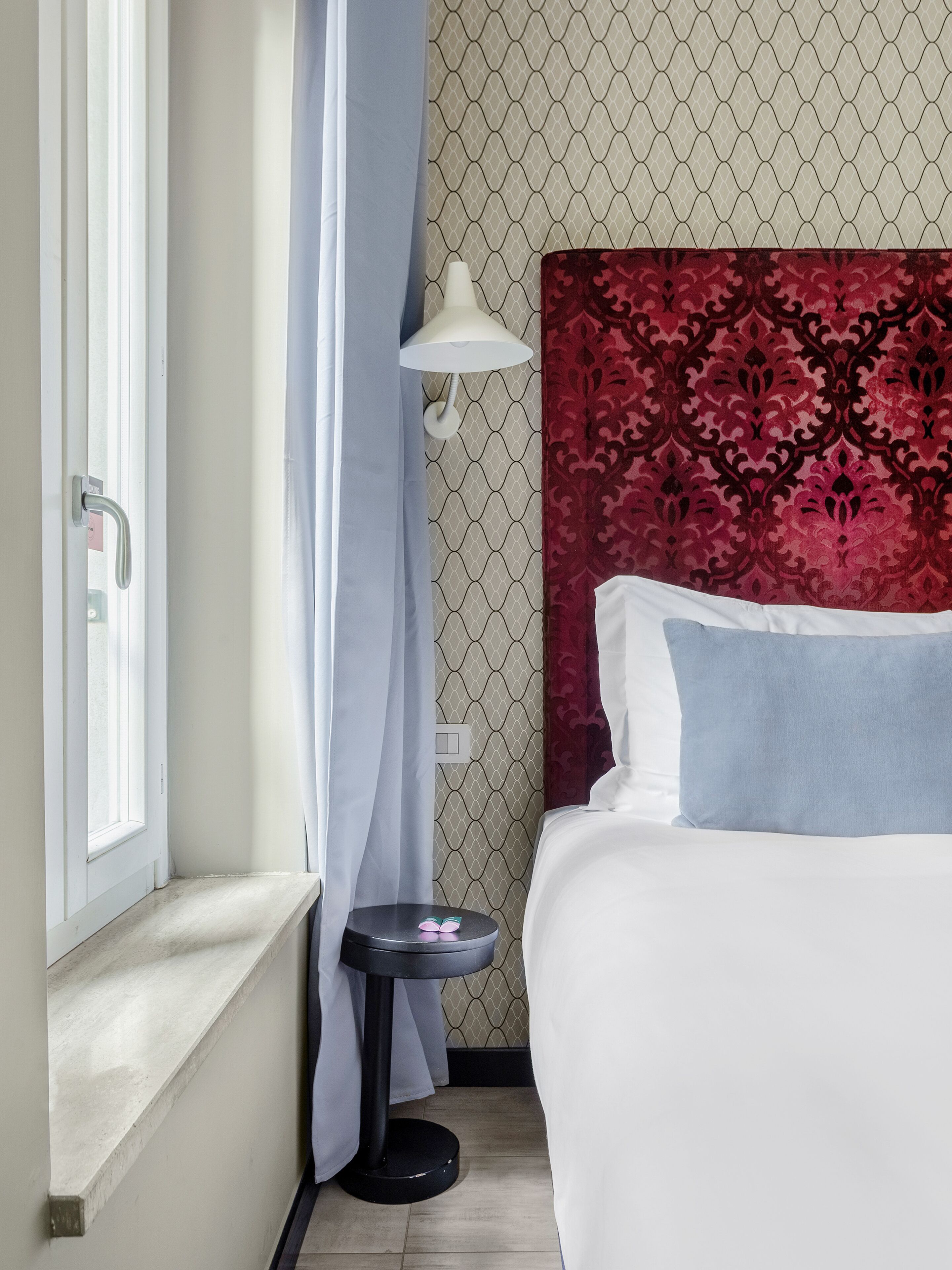 Quarto de hotel com cama, mesa de cabeceira, luminária e janela com cortinas.