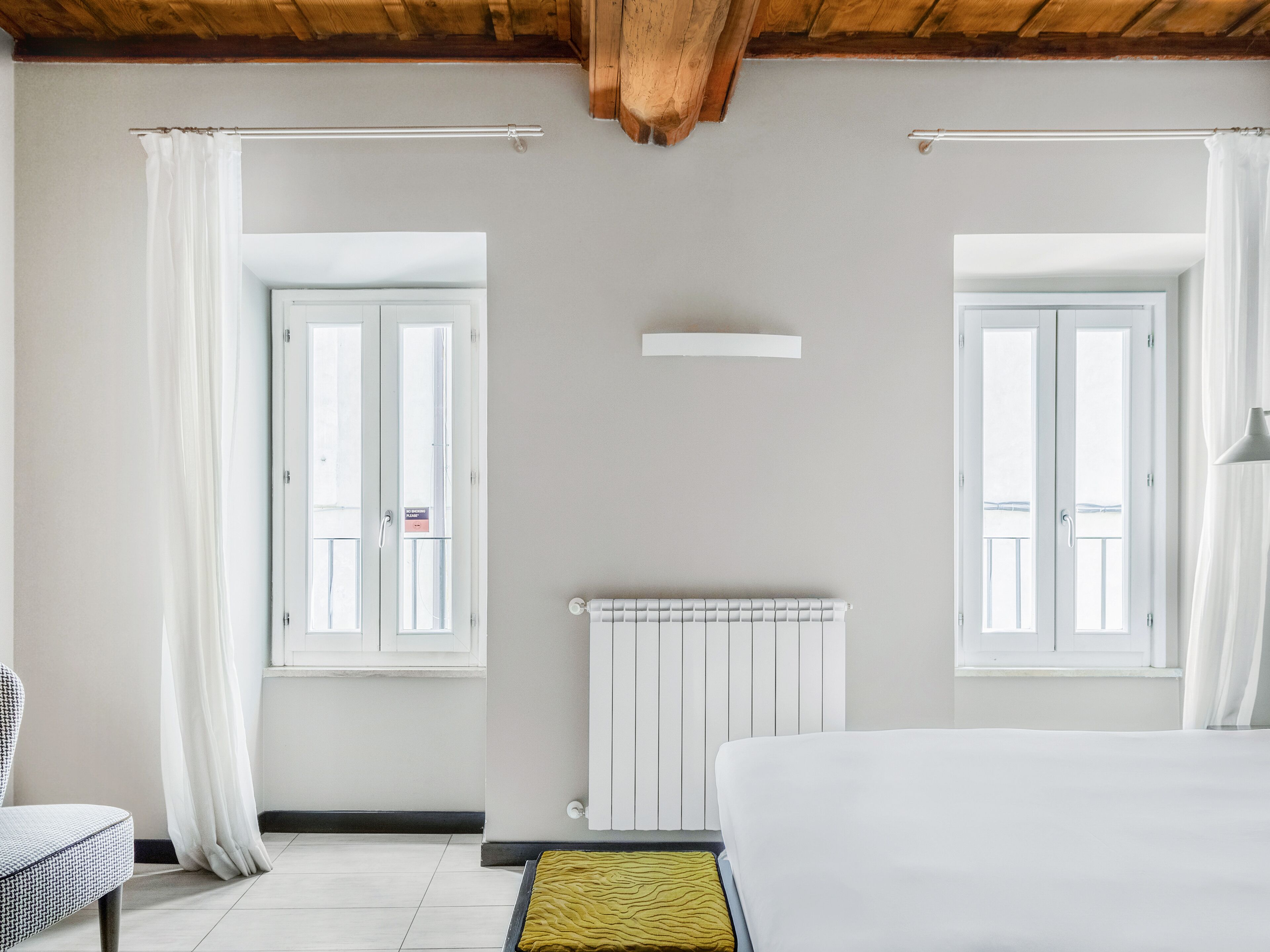 Un dormitorio con dos ventanas, un radiador, una cama y una silla.