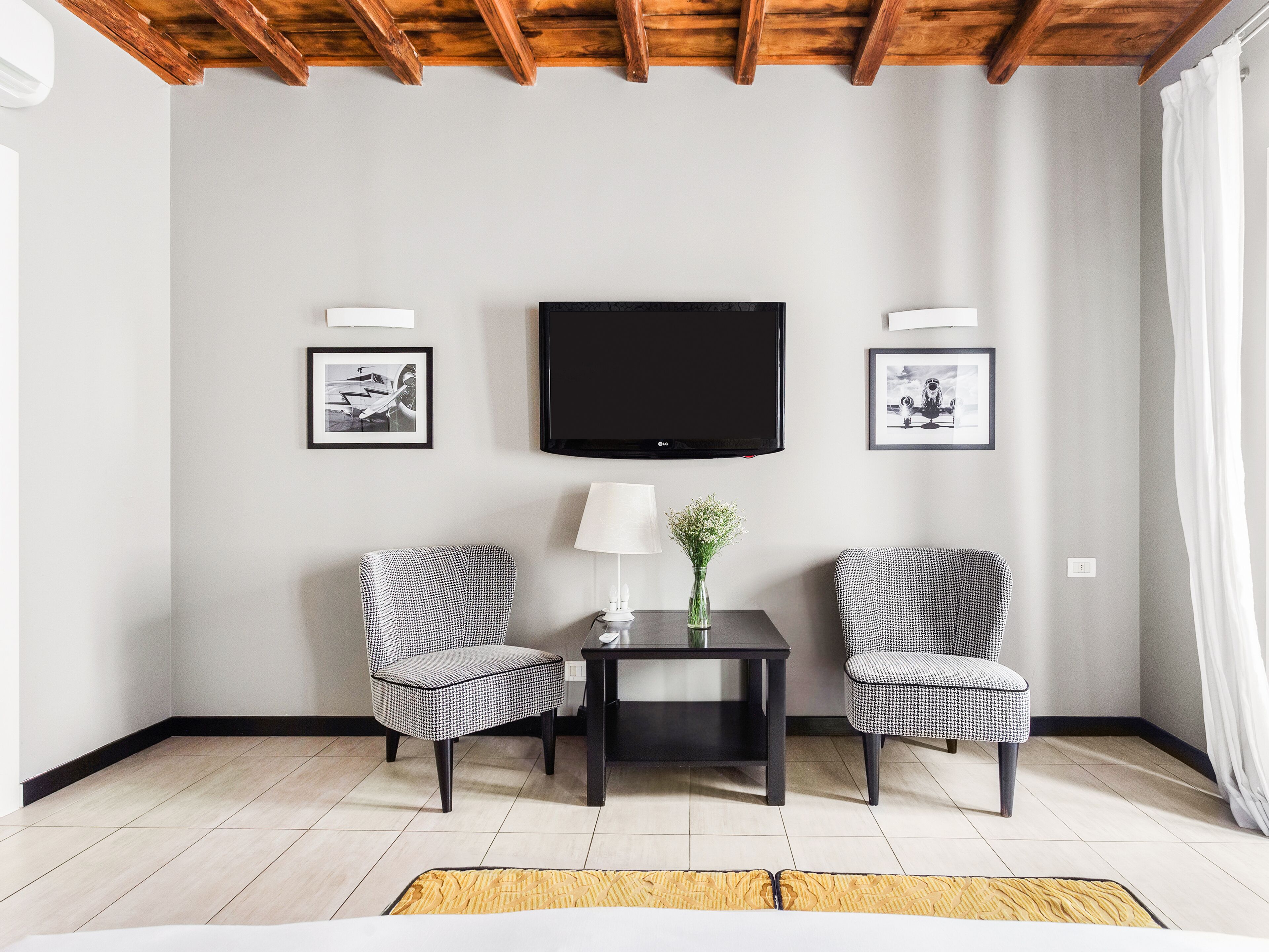 A modern living room with a flat-screen TV mounted on the wall, two patterned armchairs, a small table with a lamp and a vase, and a light-colored rug.