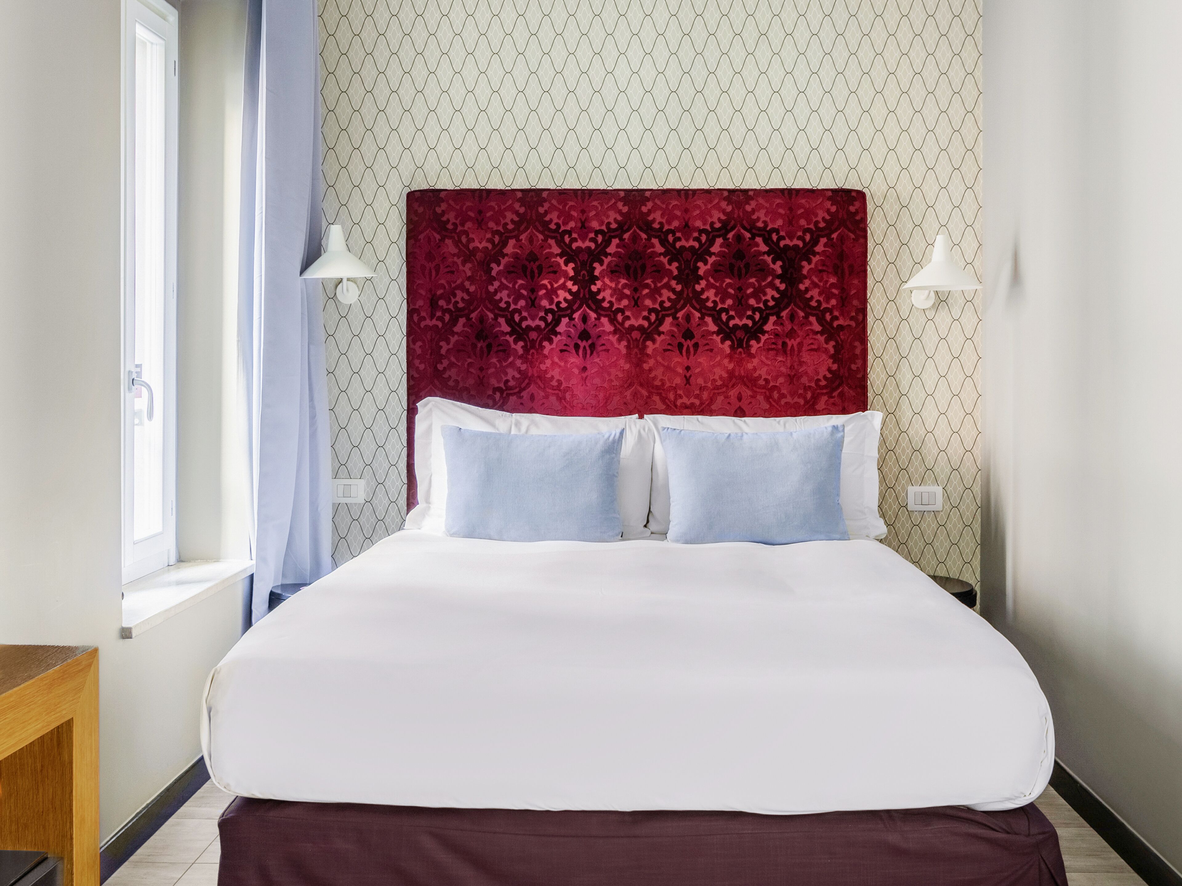 A neatly made bed with white and blue bedding, a red patterned headboard, and two bedside lamps.