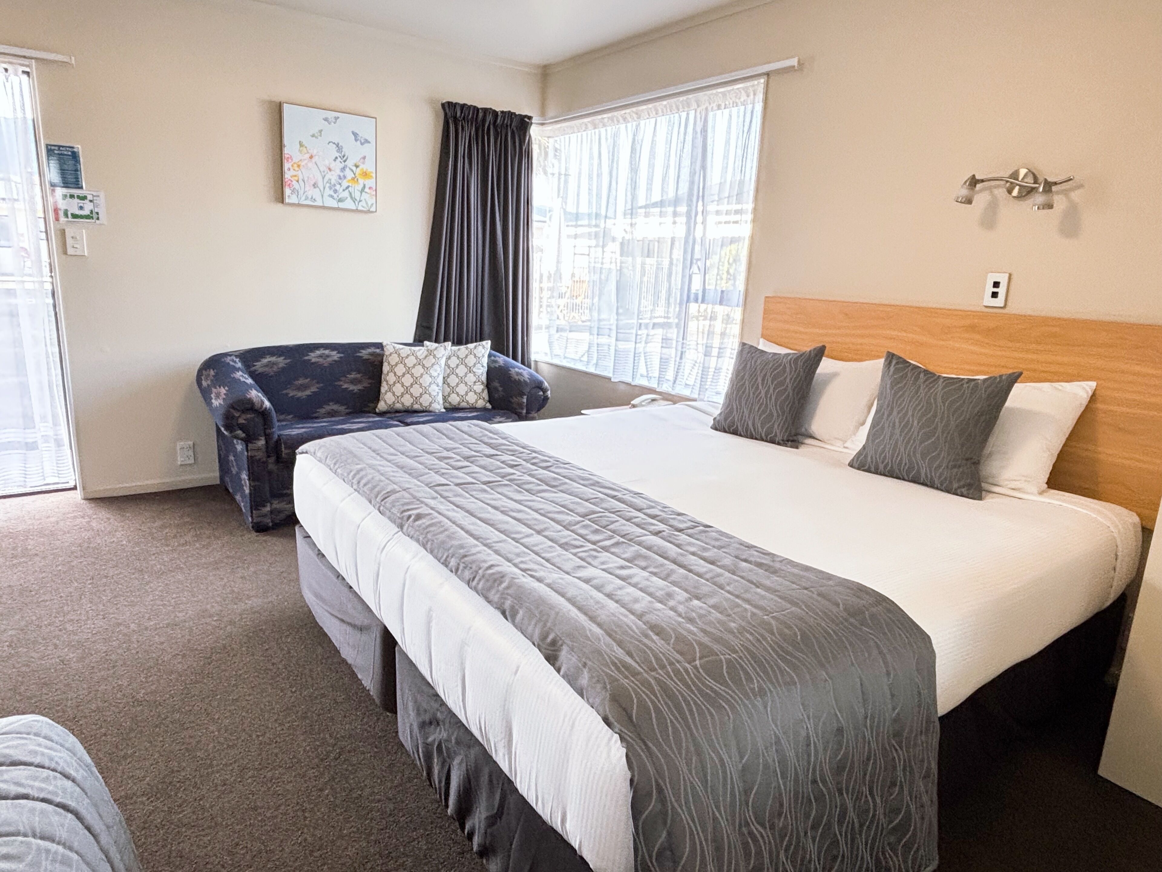 A hotel room with a large bed, a sofa, a window with curtains, and a wall-mounted light fixture.