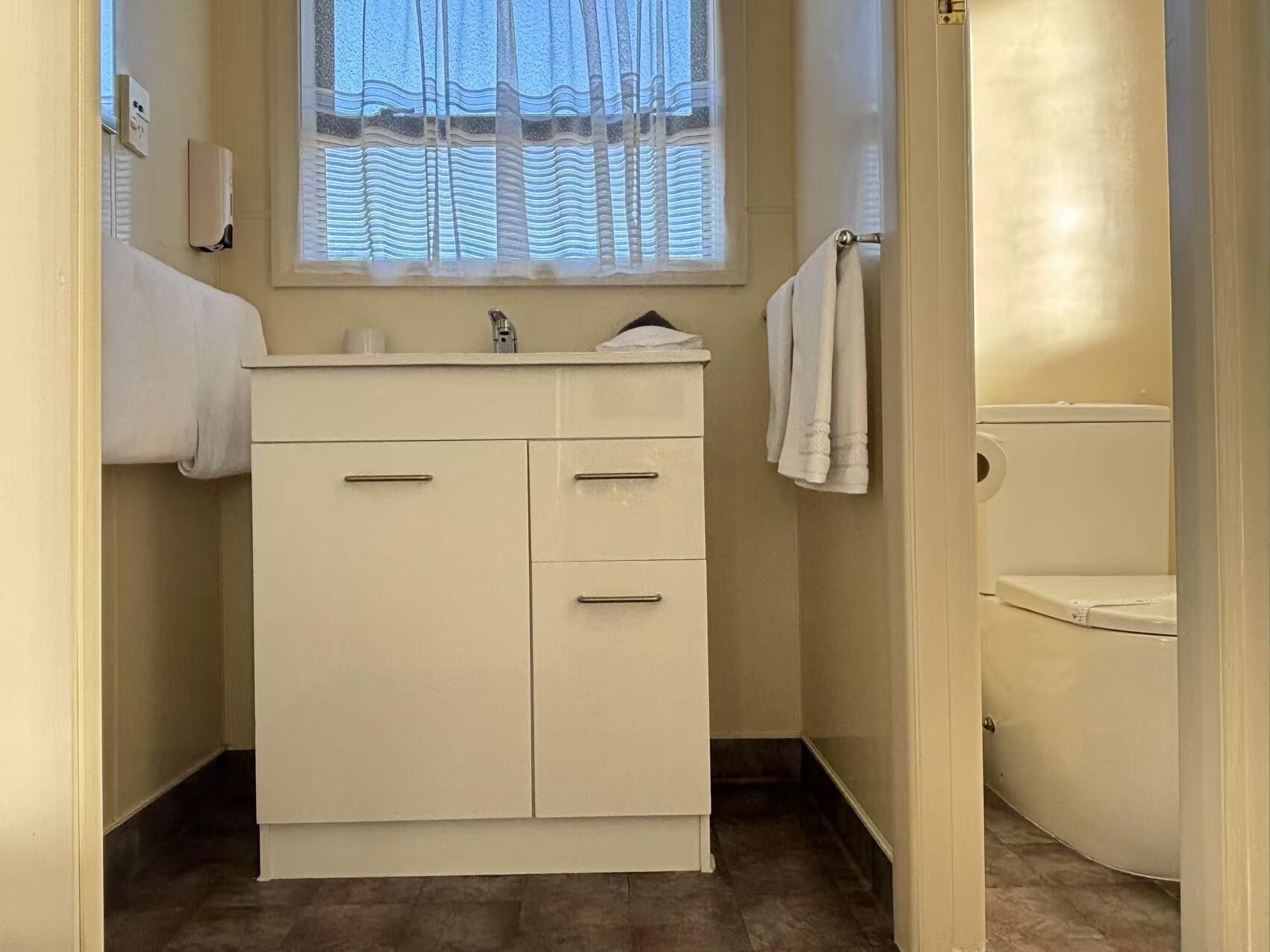 Un baño con lavamanos blanco, espejo y retrete. Se ve una ventana con cortina ligera y un perchero para toallas.