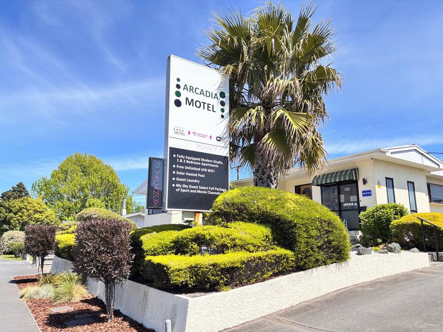 The exterior of the Arcadia Motel, featuring a large signboard with amenities listed, modern architecture, and well-maintained landscaping.