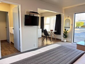A hotel room with a bed, a TV mounted on the wall, a dining area with a table and chairs, and a view of the outside.