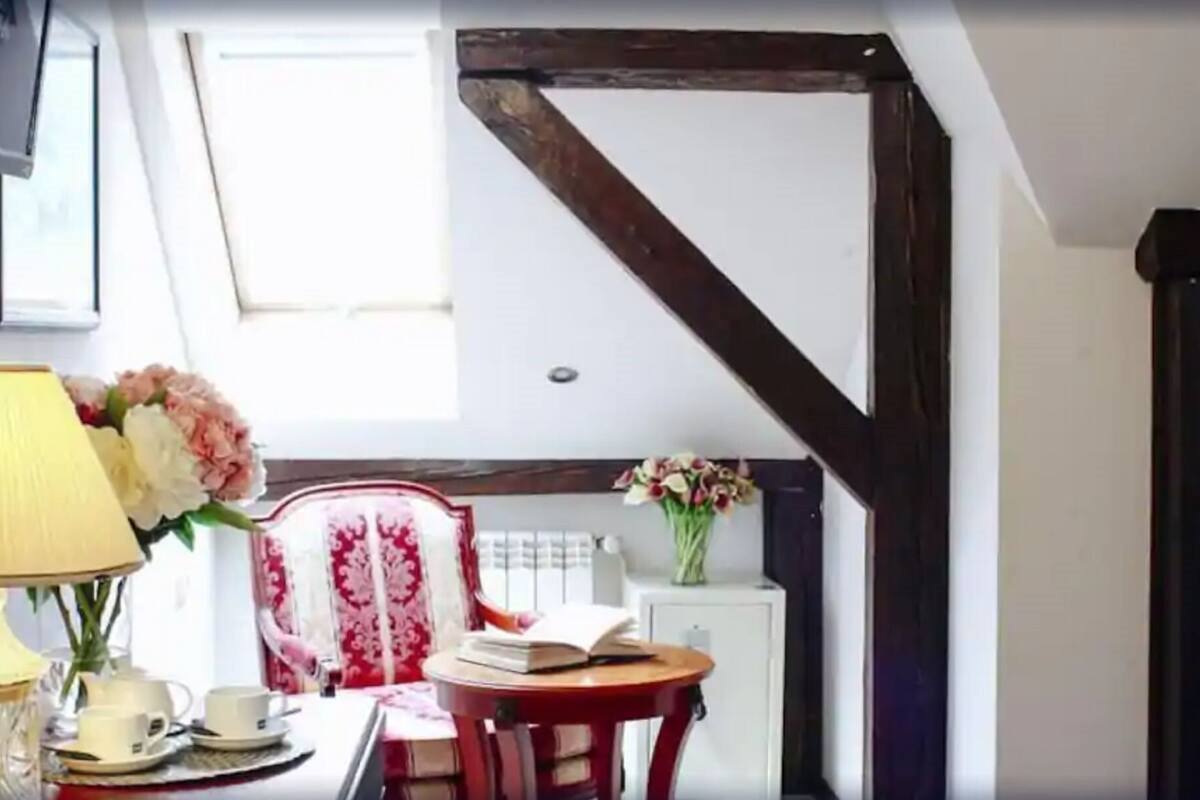 Una habitación con una viga de madera en el techo, una silla roja con estampado floral, una mesa con lámpara, un florero y un radiador.