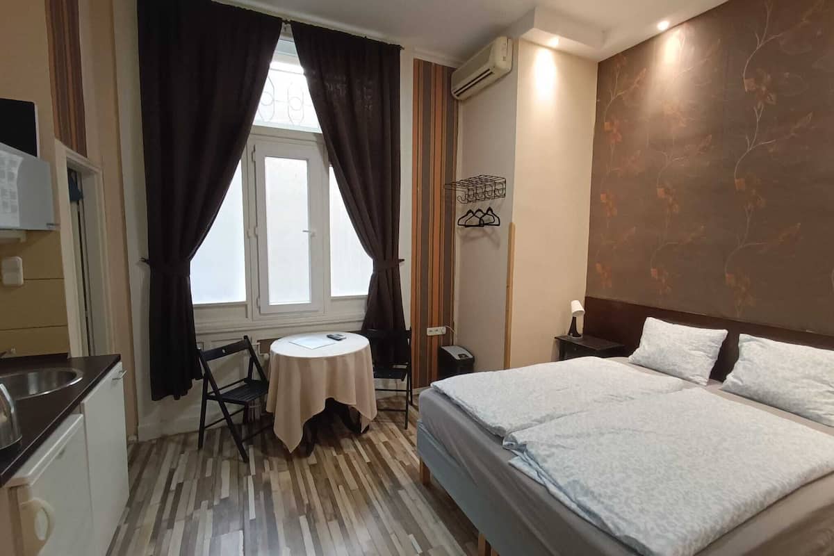 Habitación de hotel con cama, una mesita, una silla, escritorio con lavabo y ventana con cortinas.