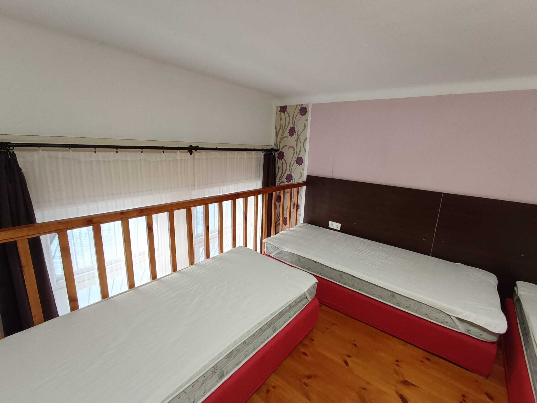 A small room with two single beds, a wooden floor, a window with curtains, and a wall with a floral pattern.
