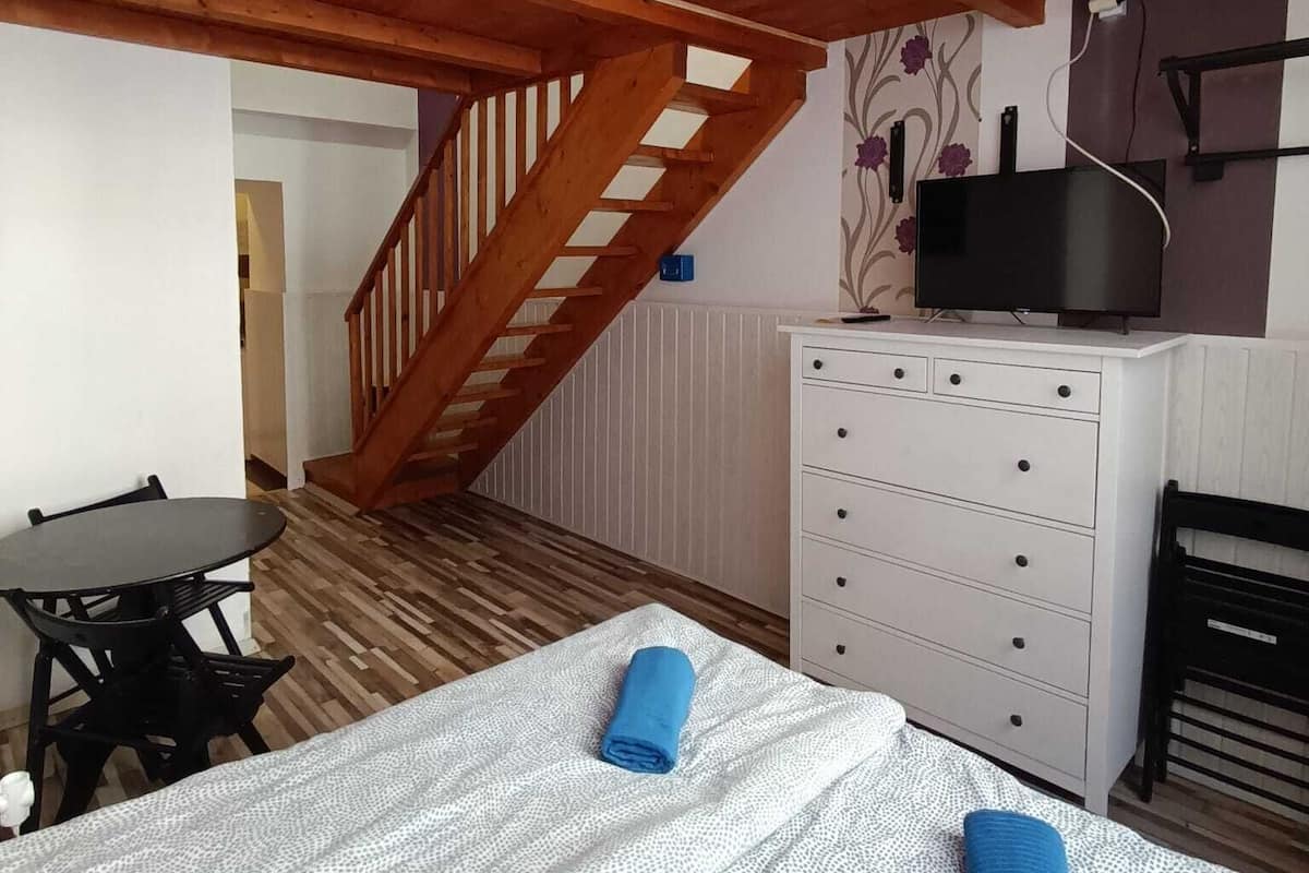 Un dormitorio con techo de madera, una cama con colcha estampada, una toalla azul, un aparador blanco, un televisor sobre un mueble bajo y una escalera.