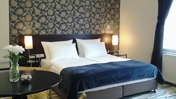A hotel room with a bed, two bedside lamps, a small table with a vase and a decorative item, and a floral-patterned wall.