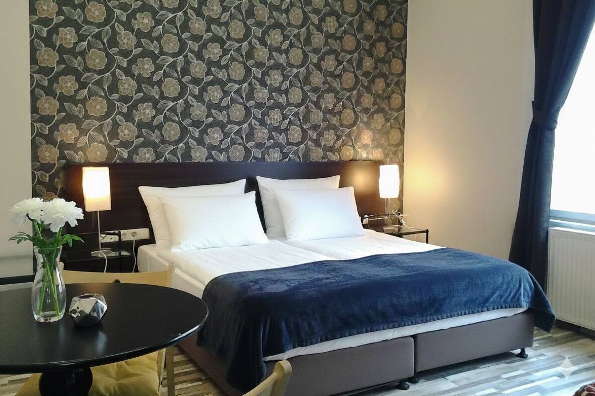 Habitación de hotel con cama, dos lámparas de noche, una mesita con un florero y un adorno, y un papel tapiz estampado con motivos florales.