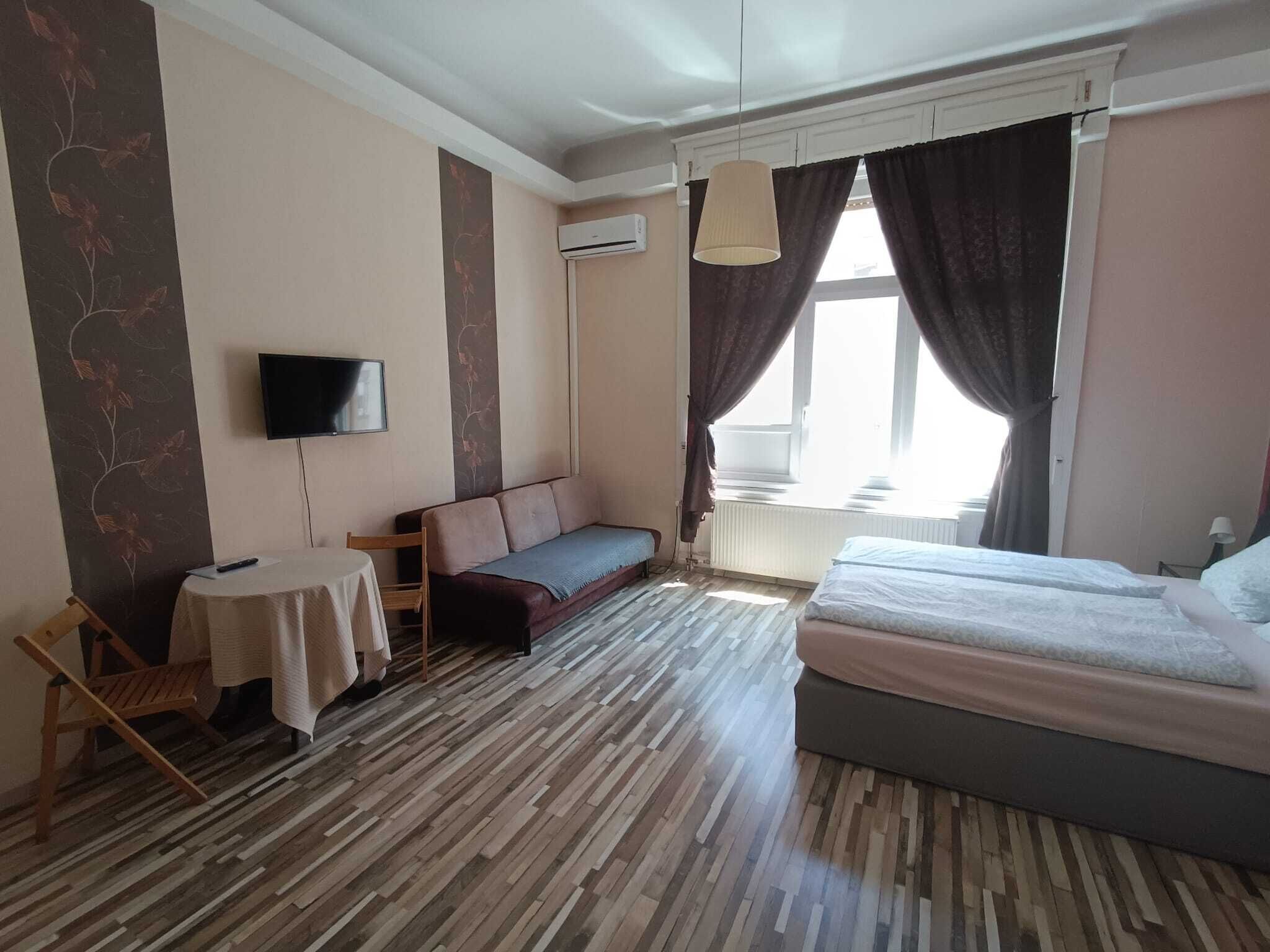 A hotel room with a bed, a sofa, a small table, a TV, and a window with curtains.