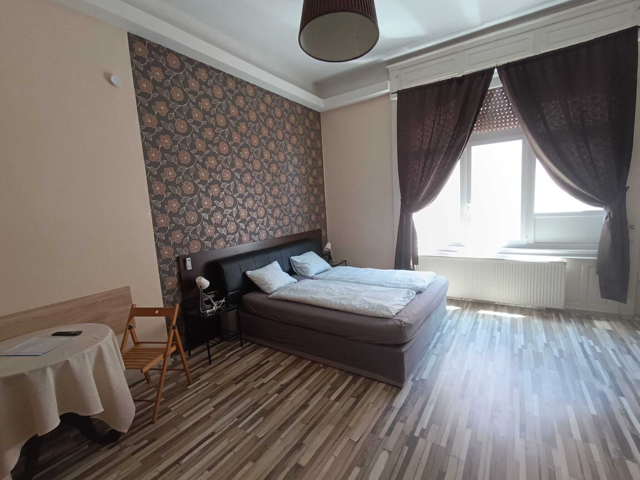 A bedroom with a bed, a small table, a chair, a radiator, and a window with curtains.