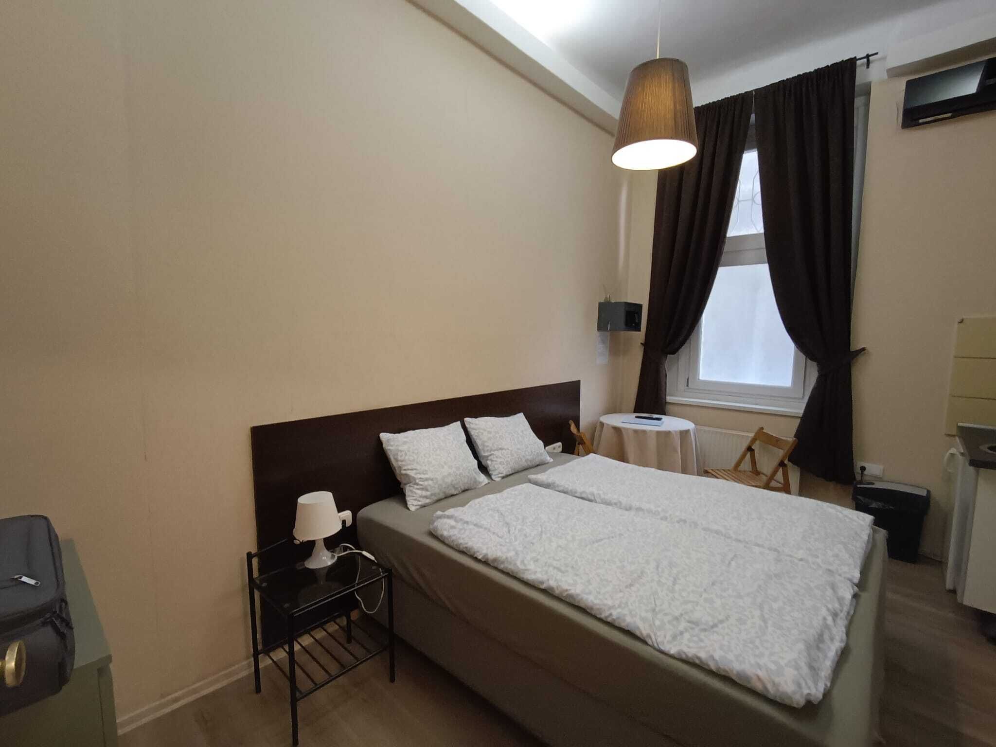 A hotel room with a single bed, a bedside table with a lamp, a small table with a chair, a window with curtains, and a TV mounted on the wall.