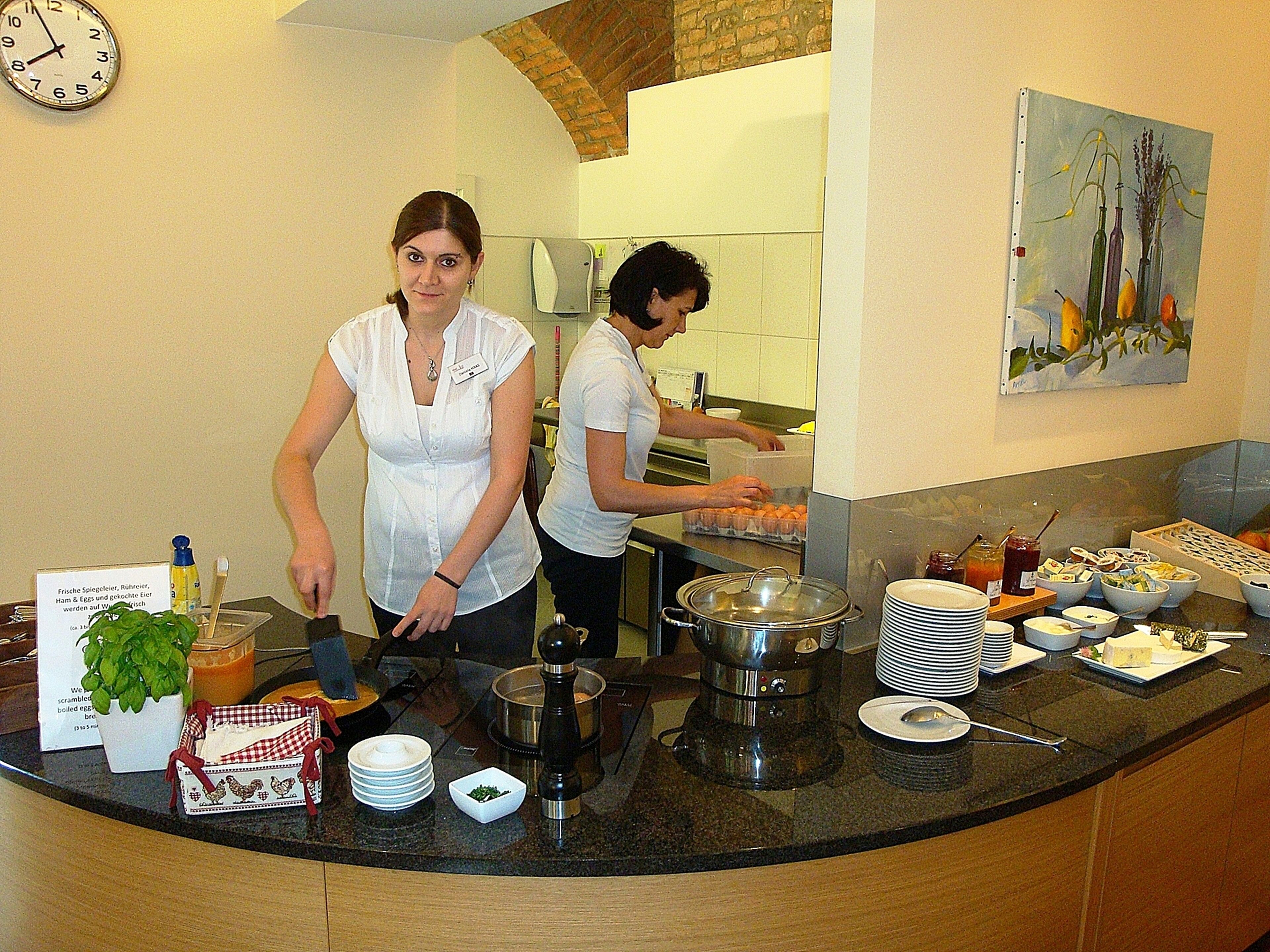 A kitchen with two people preparing food, a clock on the wall, and various kitchen items on the counter.