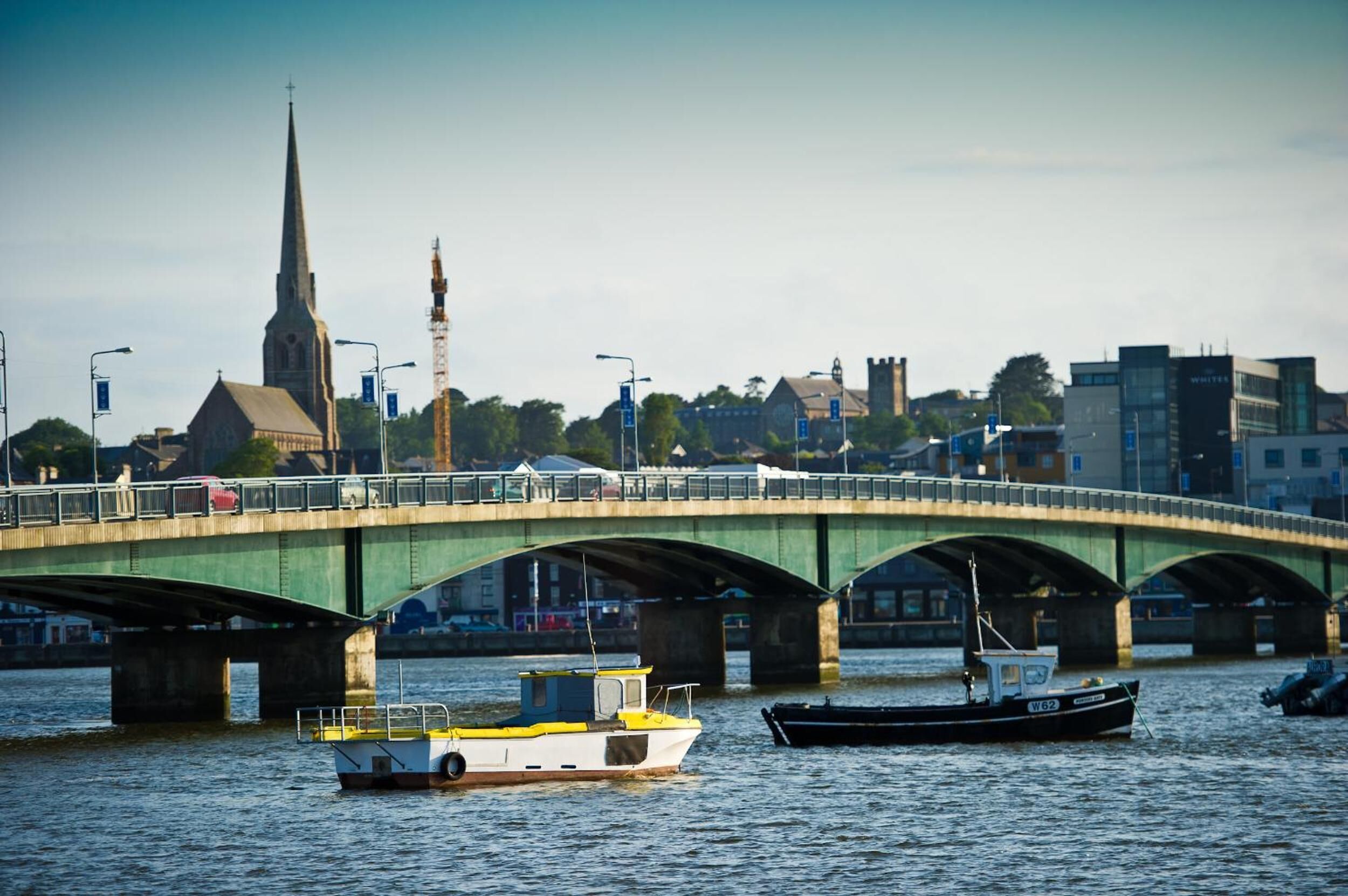 A bridge over water with boats, a church spire, and buildings in the background.