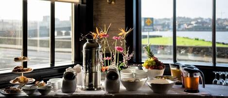 Un bufé de desayuno con diversos alimentos y bebidas, una cafetera y un jarrón con flores.