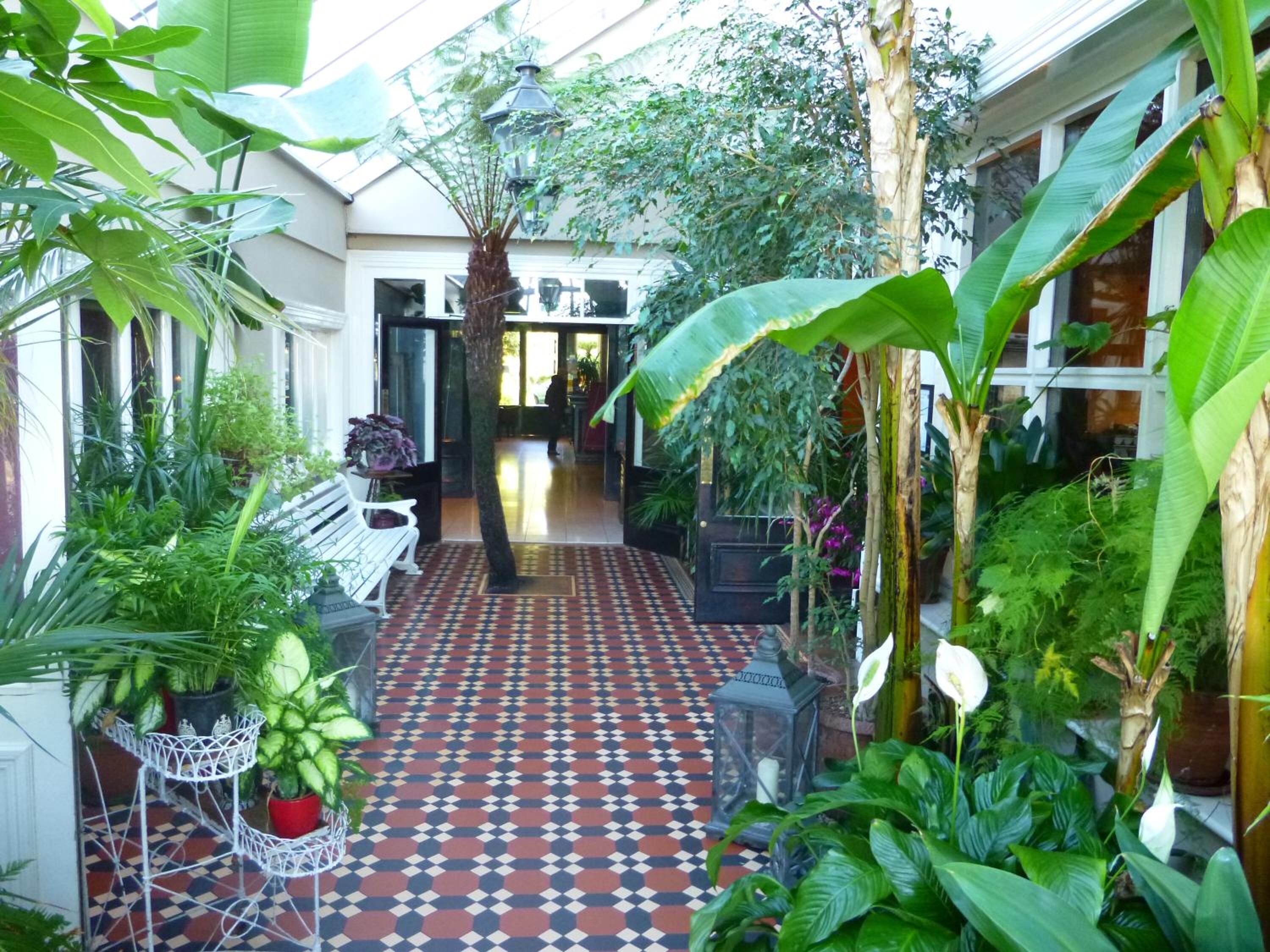 A narrow indoor corridor with a patterned floor, surrounded by various potted plants and a bench.