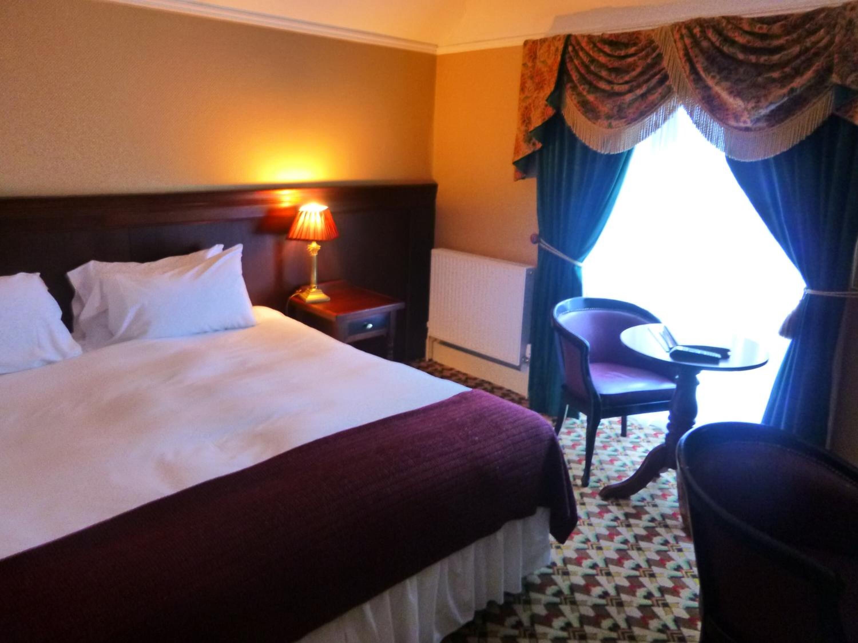 A hotel room with a large bed, a bedside table with a lamp, a chair, and a view of the outdoors through a window with curtains.