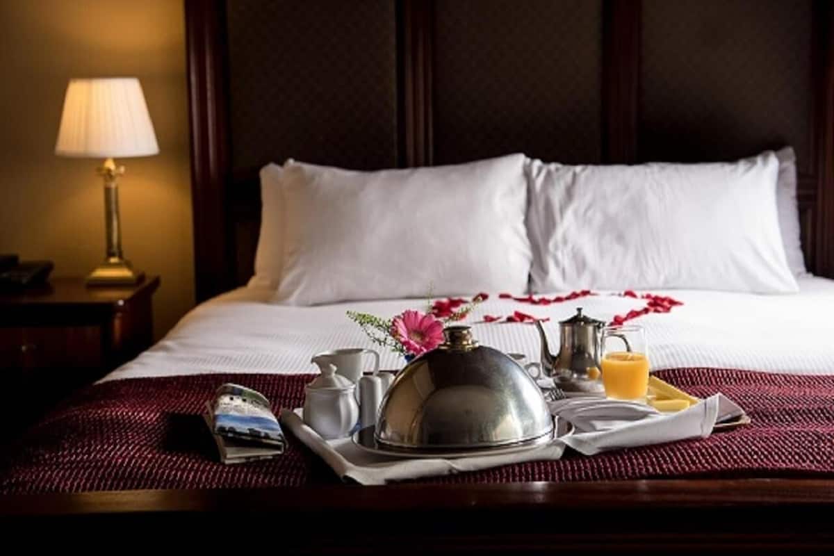 Ein ordentlich bezogenes Bett mit weißen Leinen, ein Tablett mit Frühstücksartikeln, ein Saftkrug mit Orangensaft und ein kleiner Vasen mit rosa Blumen.