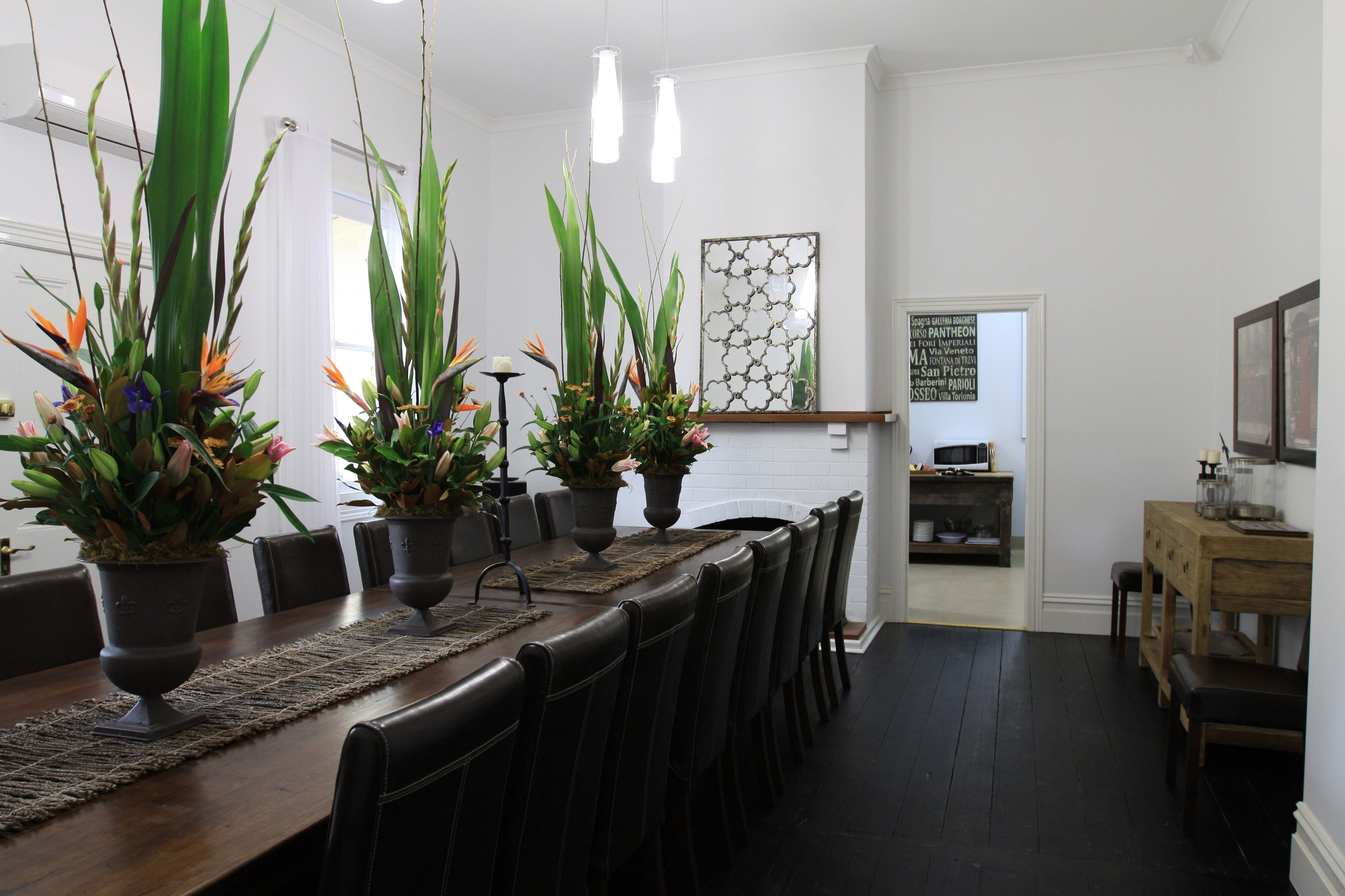A dining area with a long wooden table, black chairs, and potted plants. There is a kitchen visible in the background.