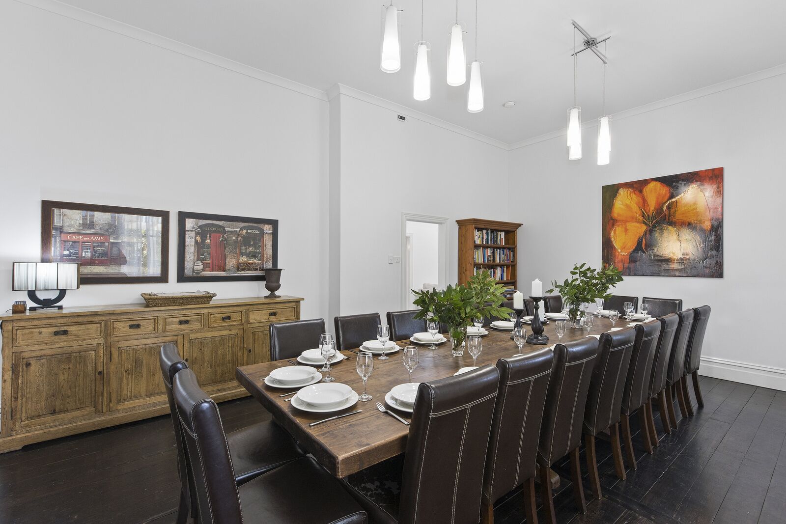 Une salle à manger avec une table en bois dressée pour six personnes, entourée de chaises en cuir foncé. Au-dessus de la table, on trouve des lampes suspendues, un buffet en bois avec un miroir, et deux tableaux encadrés accrochés au mur.