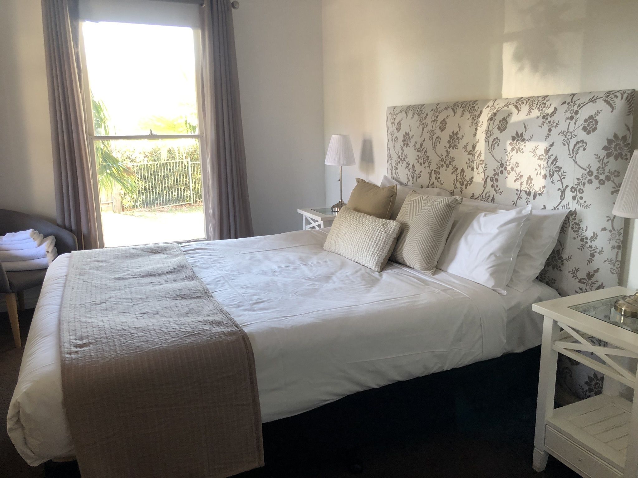 A neatly made bed with a white comforter and several pillows, a bedside table with a lamp, and a window with curtains.
