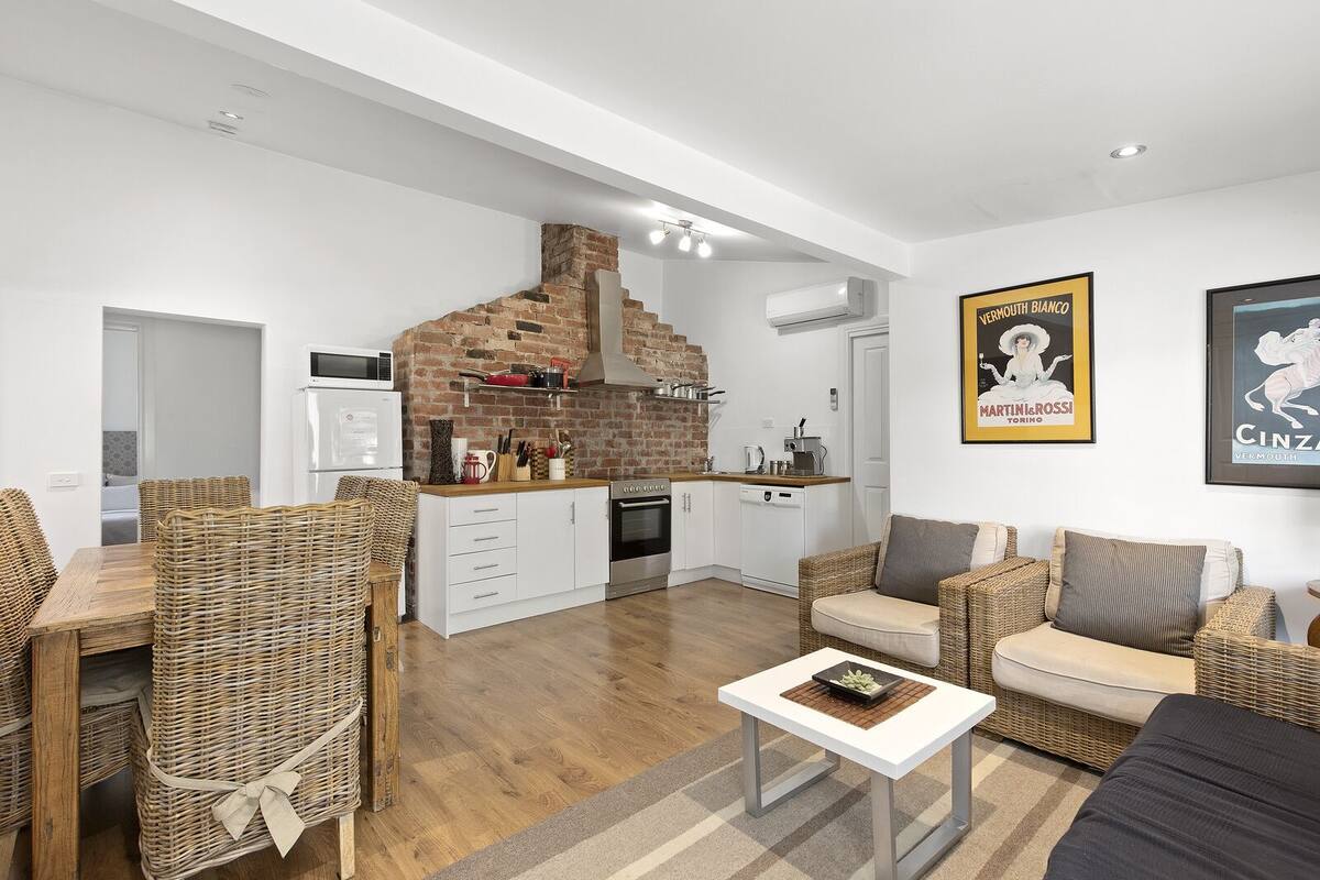 A modern kitchen with a brick wall, white cabinetry, and a built-in oven. There is a dining area with wicker furniture and a coffee table.