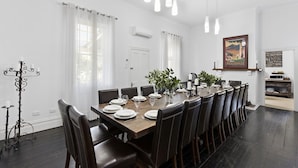 Een eetkamer met een lange tafel gedekt voor zes personen, omringd door donkere lederen stoelen. Er zijn witte gordijnen bij de ramen, een kandelaar links en een lijst met een afbeelding aan de muur.