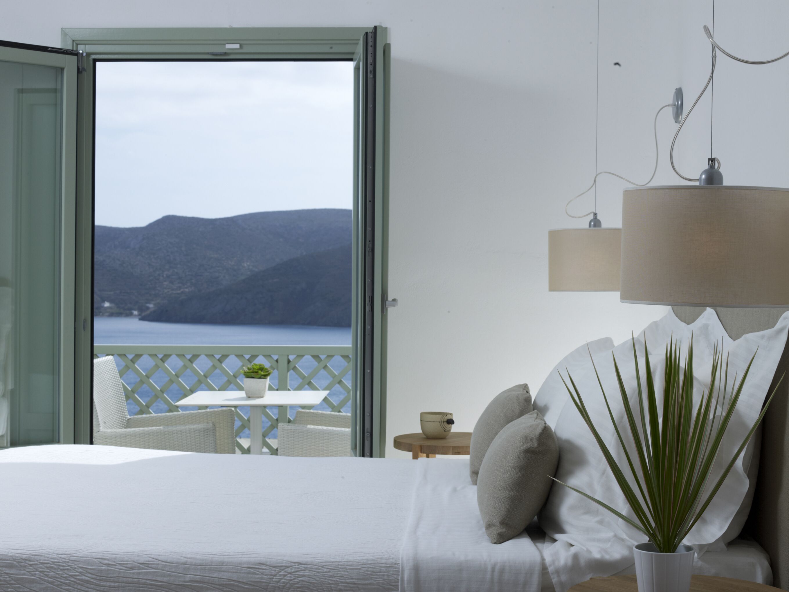 A bedroom with a bed, a potted plant, a nightstand, a table with a bowl, and a view of the sea and mountains through an open door.