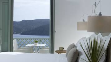 A bedroom with a bed, a potted plant, a nightstand, a table with a bowl, and a view of the sea and mountains through an open door.