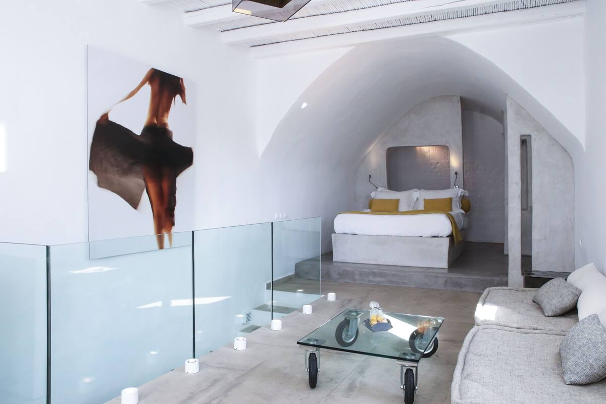 Une chambre d’hôtel moderne avec une table basse en verre, un canapé, un lit doté d’une tête de lit blanche et jaune, et un grand tableau abstrait accroché au mur.