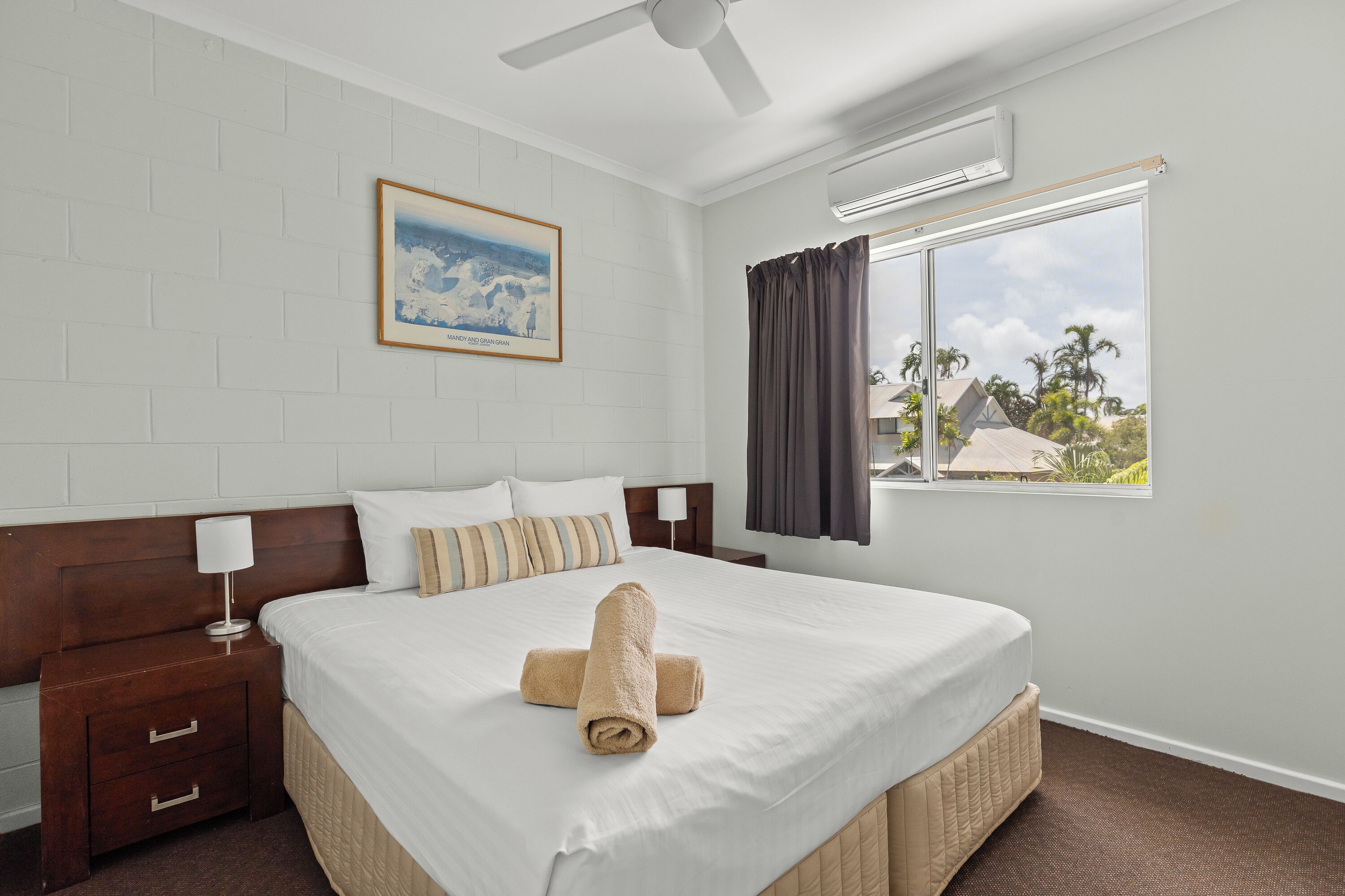 A hotel room with a bed, a nightstand, a ceiling fan, a window with curtains, and a painting on the wall.