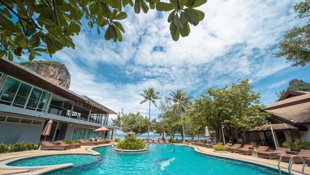 A resort pool with clear blue water, surrounded by lounge chairs and umbrellas, with a view of the ocean and a building with large windows. - Sand Sea Resort (Krabi)