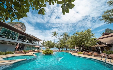 A resort pool with clear blue water, surrounded by lounge chairs and umbrellas, with a view of the ocean and a building with large windows.